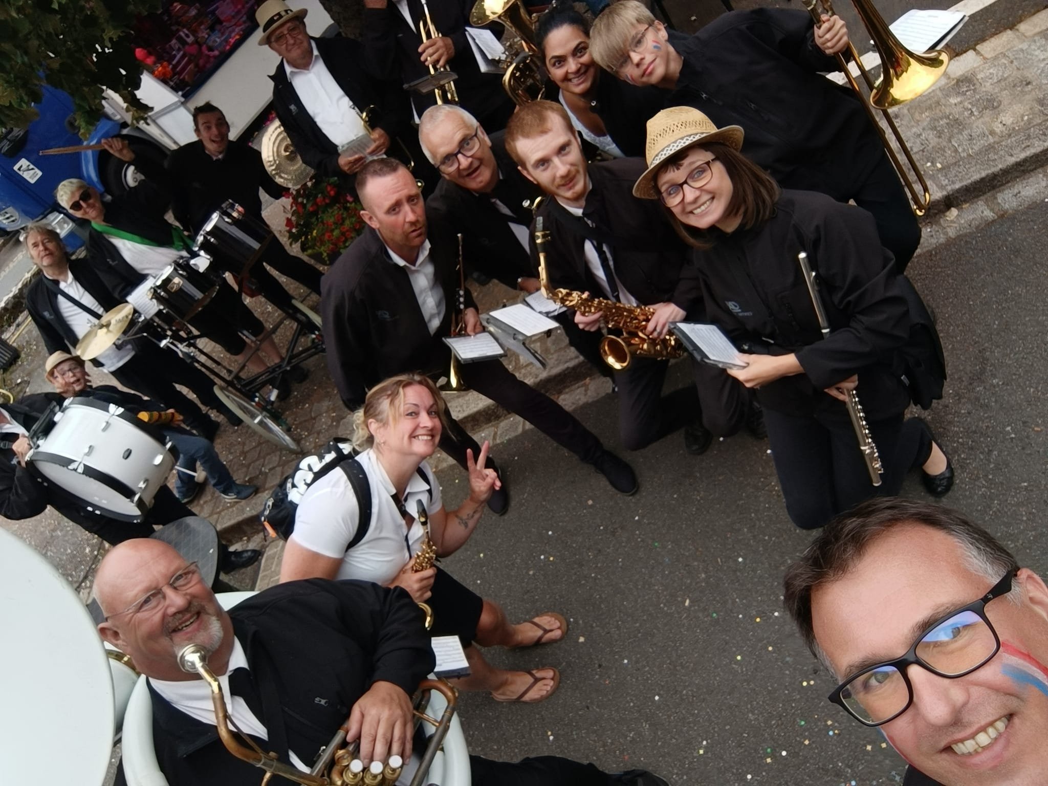 Un groupe de musiciens posant pour une photo, certains tenant des instruments de musique tels que saxophone, trombone, batterie et autres, à l'extérieur.