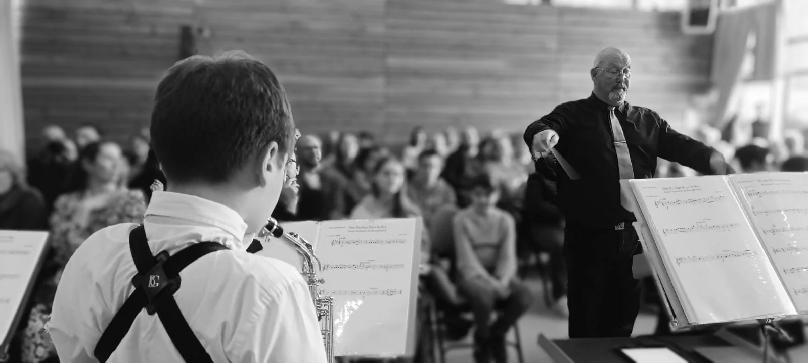 Un jeune musicien joue du saxophone lors d'un concert avec un chef d'orchestre dirigeant en face. L'auditoire est composé de plusieurs personnes assises dans une salle adjacente en arrière-plan.