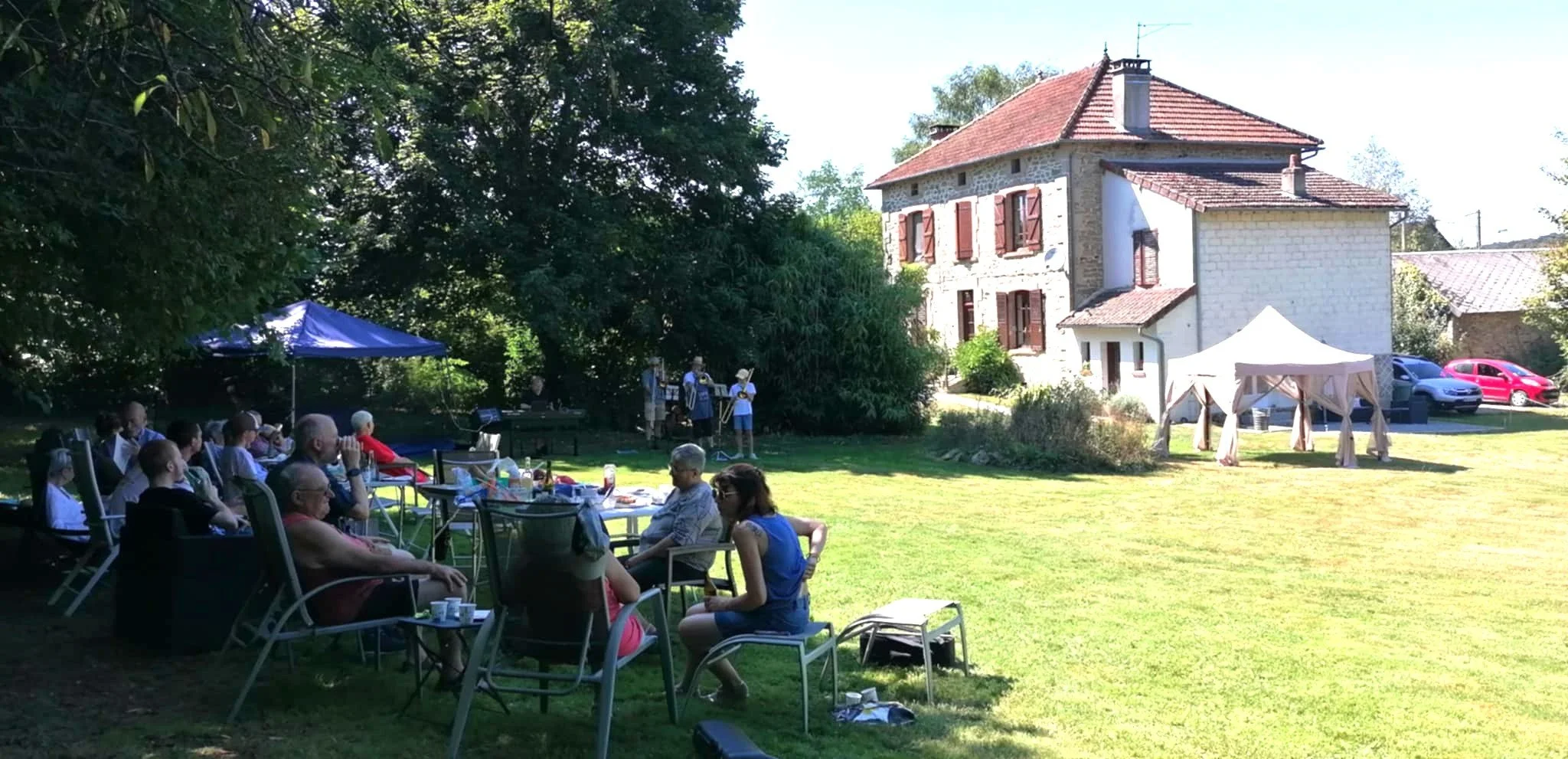 Groupe de personnes assises sous un arbre lors d'une réunion ou d'une fête en plein air avec une maison en pierre en arrière-plan et un espace vert bien entretenu