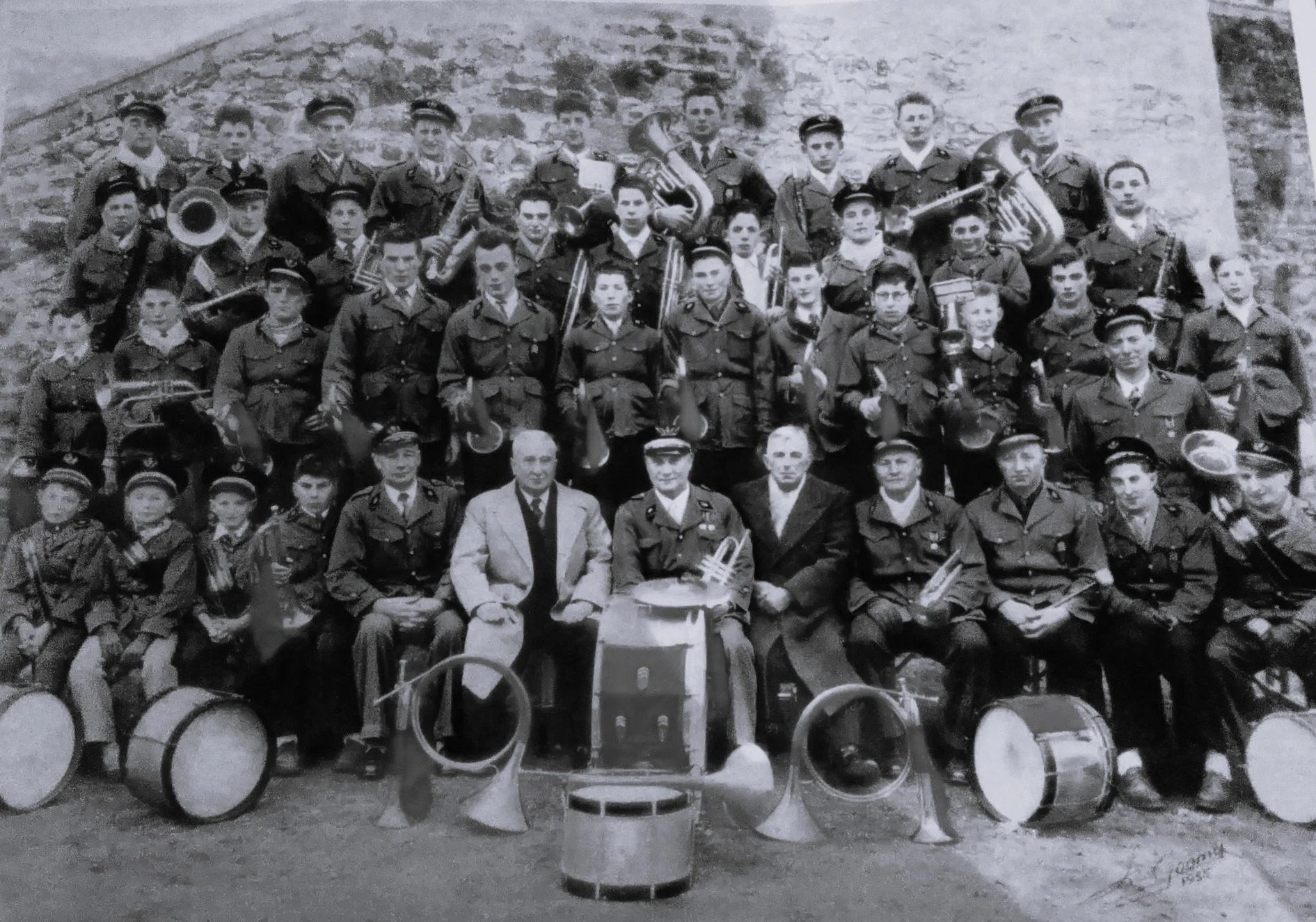 Photo en noir et blanc d’un groupe de musiciens militaires, assis et debout, avec divers instruments de musique, en extérieur devant un mur de pierre.