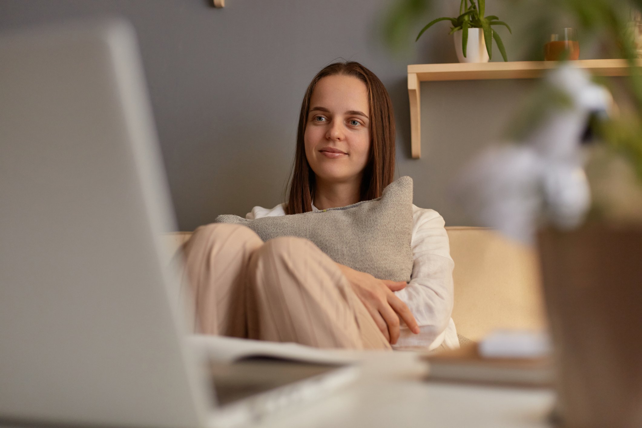 A woman sitting on a sofa, holding a pillow, with a laptop in the foreground and a shelf with a plant behind her.