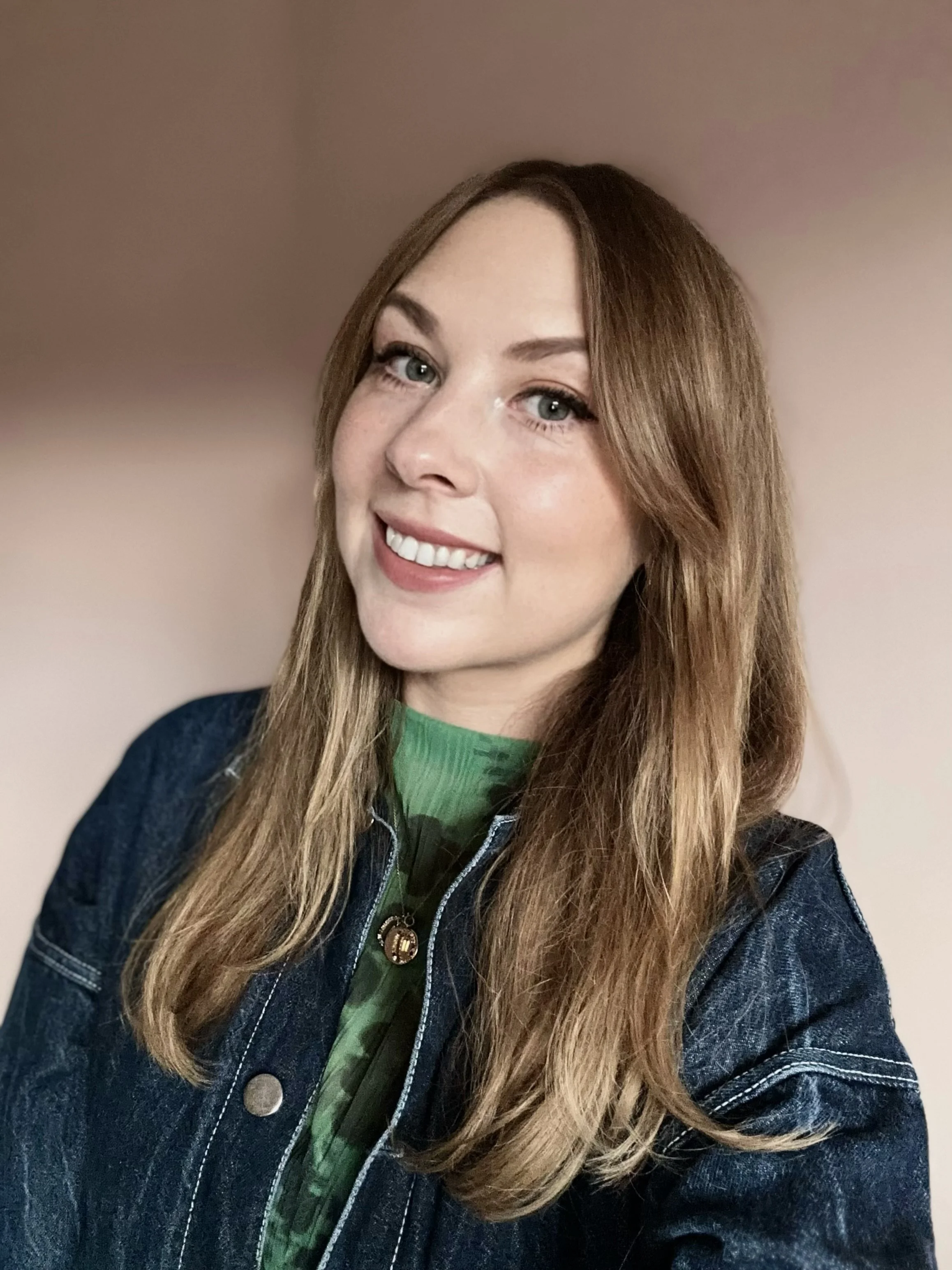 A smiling young woman with long, light brown hair, wearing a green shirt and a dark denim jacket, standing against a neutral pink background.