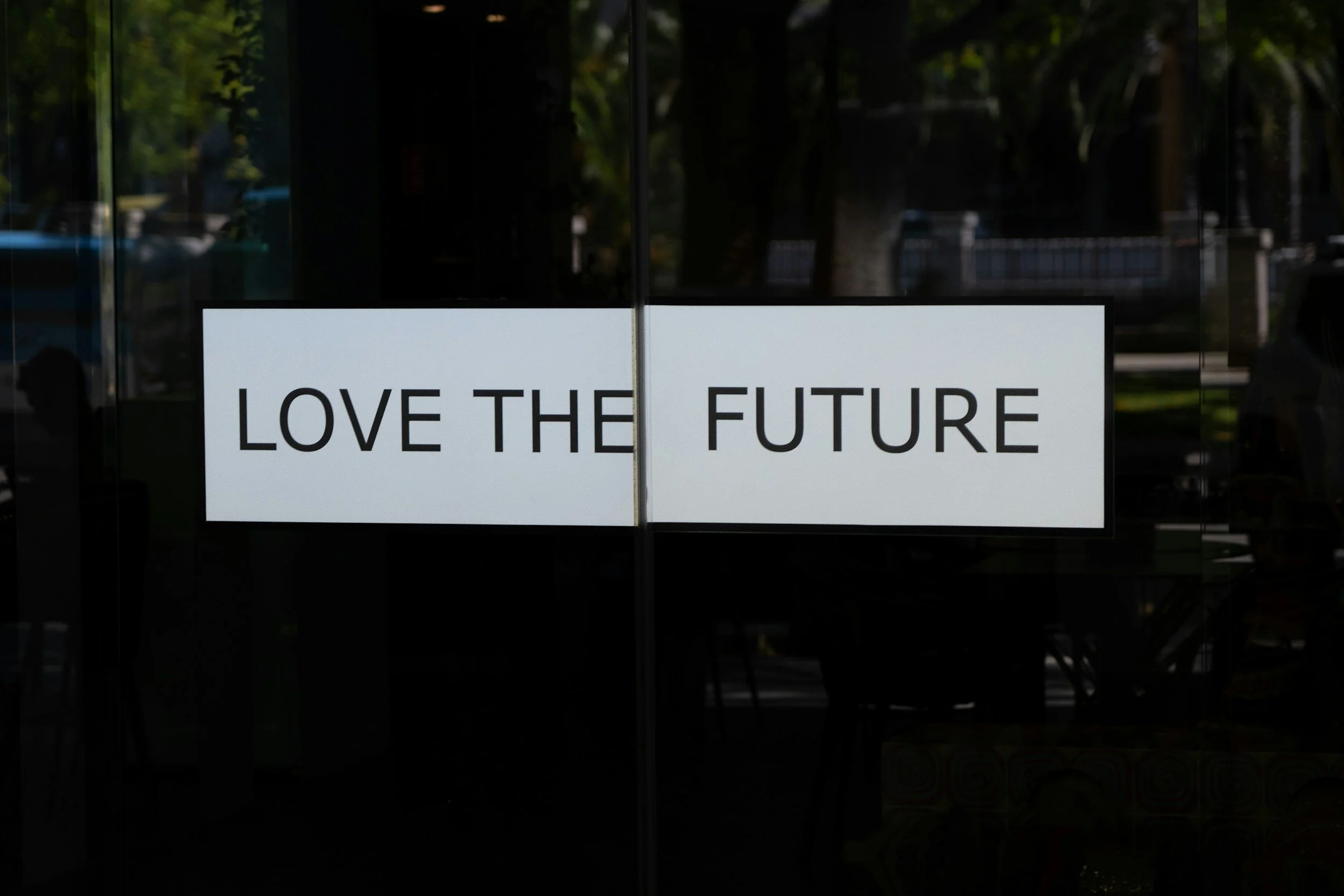 A sign on a glass door or window that reads 'LOVE THE FUTURE' with reflection of trees and outdoor scene in the background.
