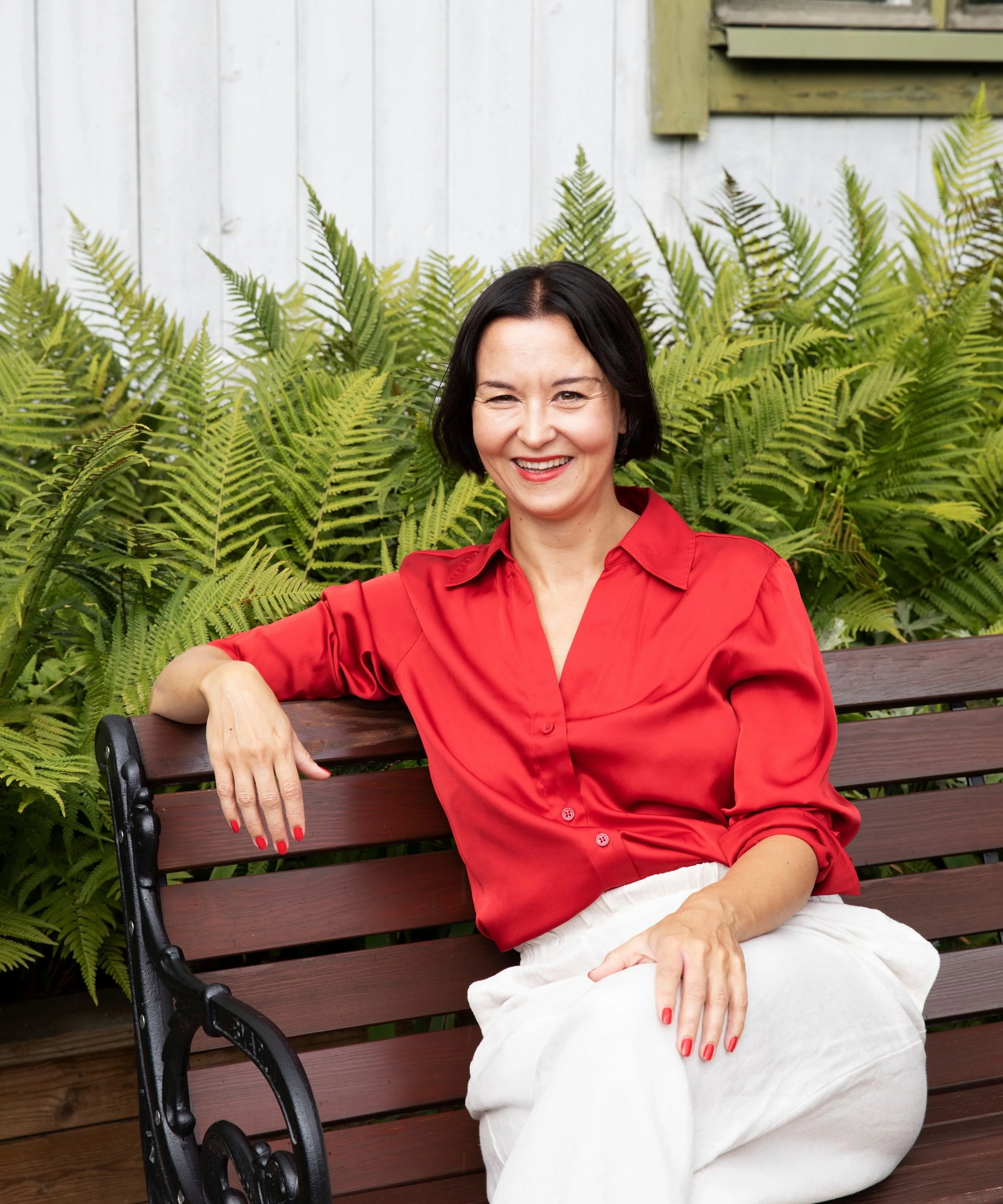 A woman with short dark hair wearing a red blouse and white pants, sitting on a wooden park bench, smiling, with green ferns and a white wall in the background.