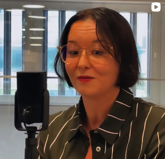 A woman with short dark hair and glasses speaking into a microphone in an office setting
