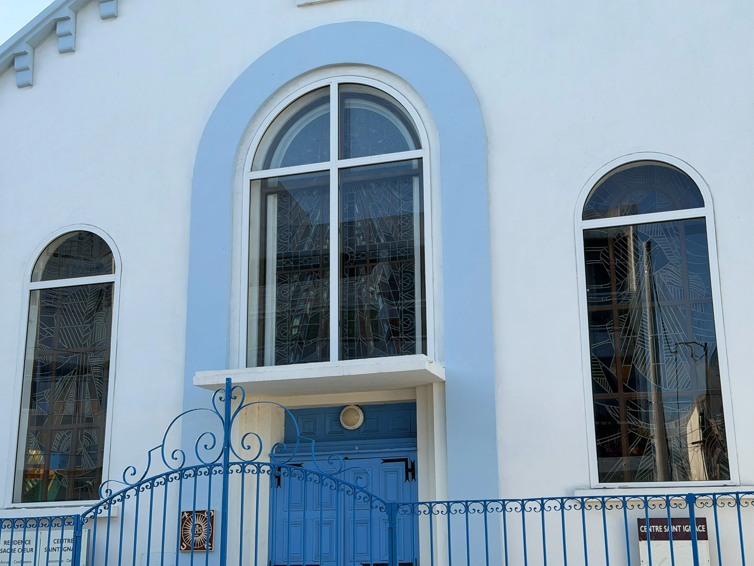 Blue church with three tall arched stained glass windows and a matching blue gate in the foreground.