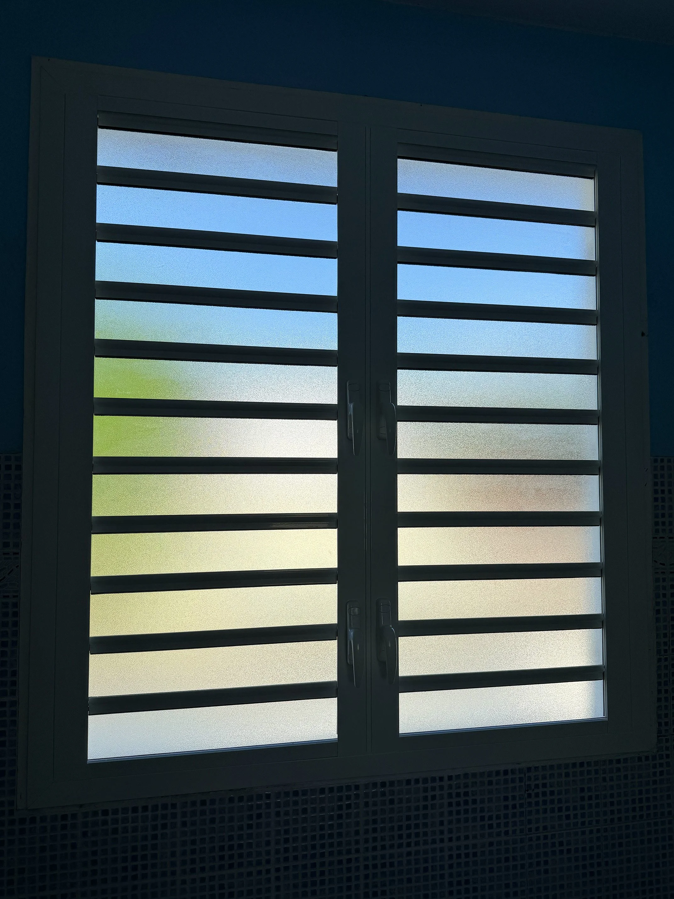 A window with white horizontal slats, showing a blue sky with some clouds outside.