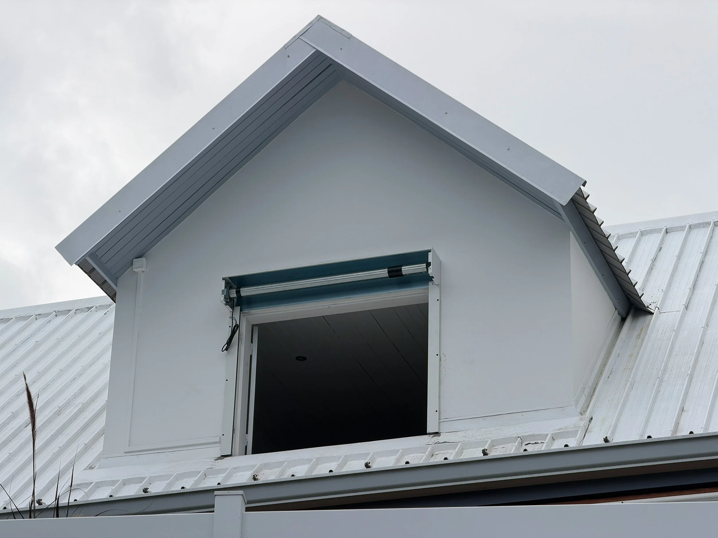 Close-up of a house's attic window opening with a partially extended metal rolling shutter, white exterior wall, and a white metal roof, under a cloudy sky.