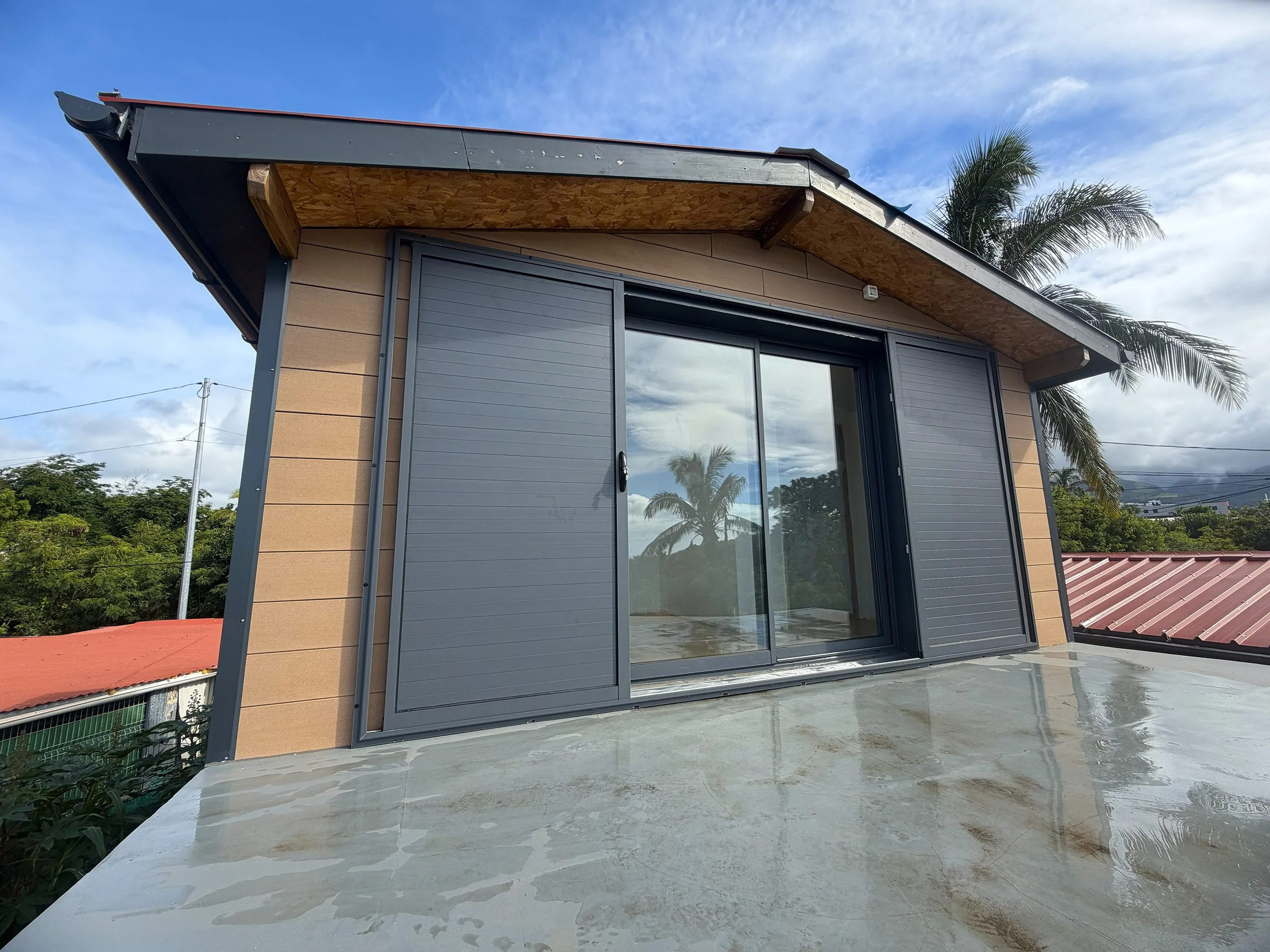 A house with a sloped roof, sliding glass doors, and mounted gray shutters, with a concrete balcony and tropical trees in the background.