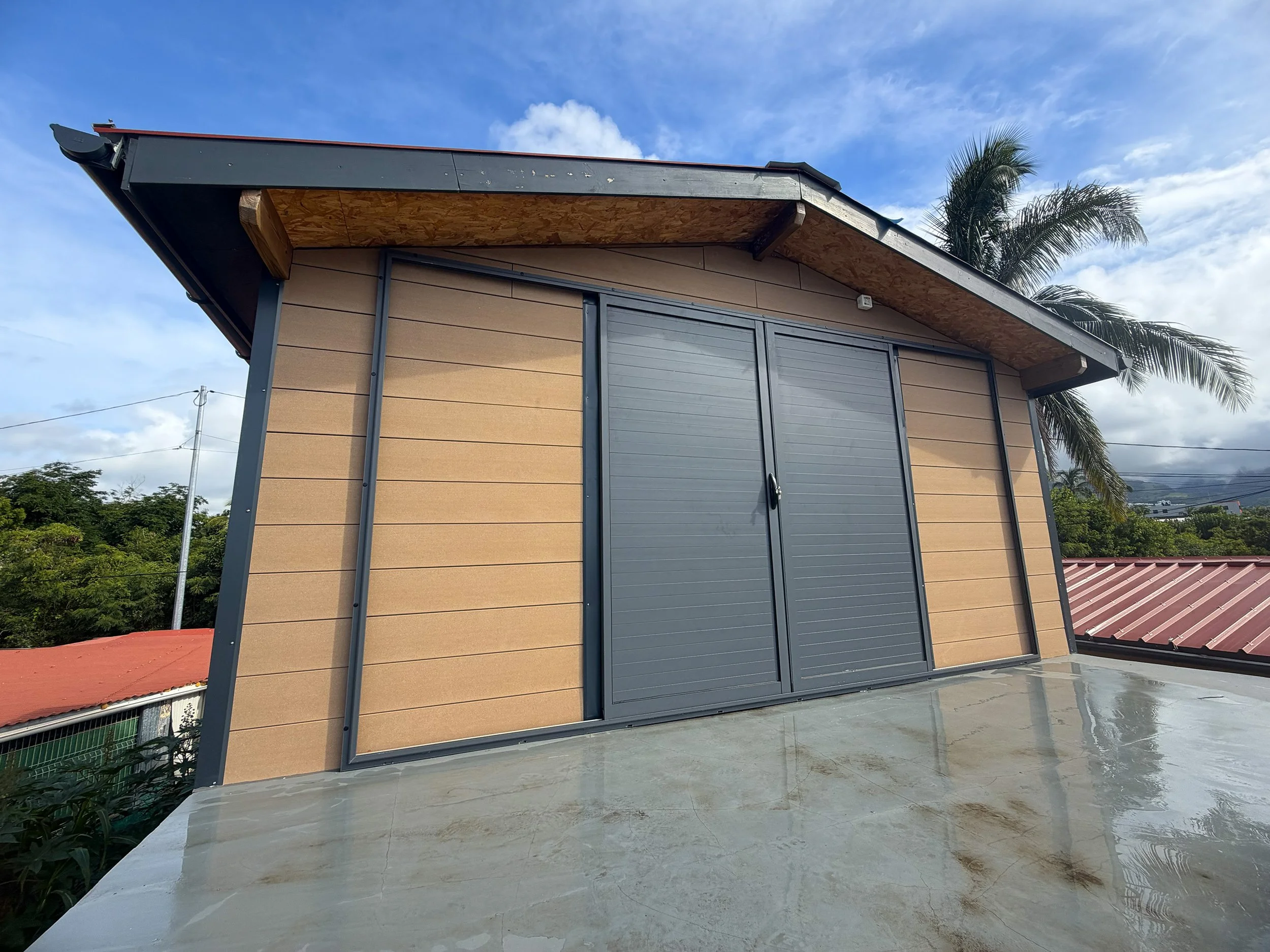 Small house with beige siding, gray sliding door, metal roof, and palm trees in the background, under a partly cloudy sky.
