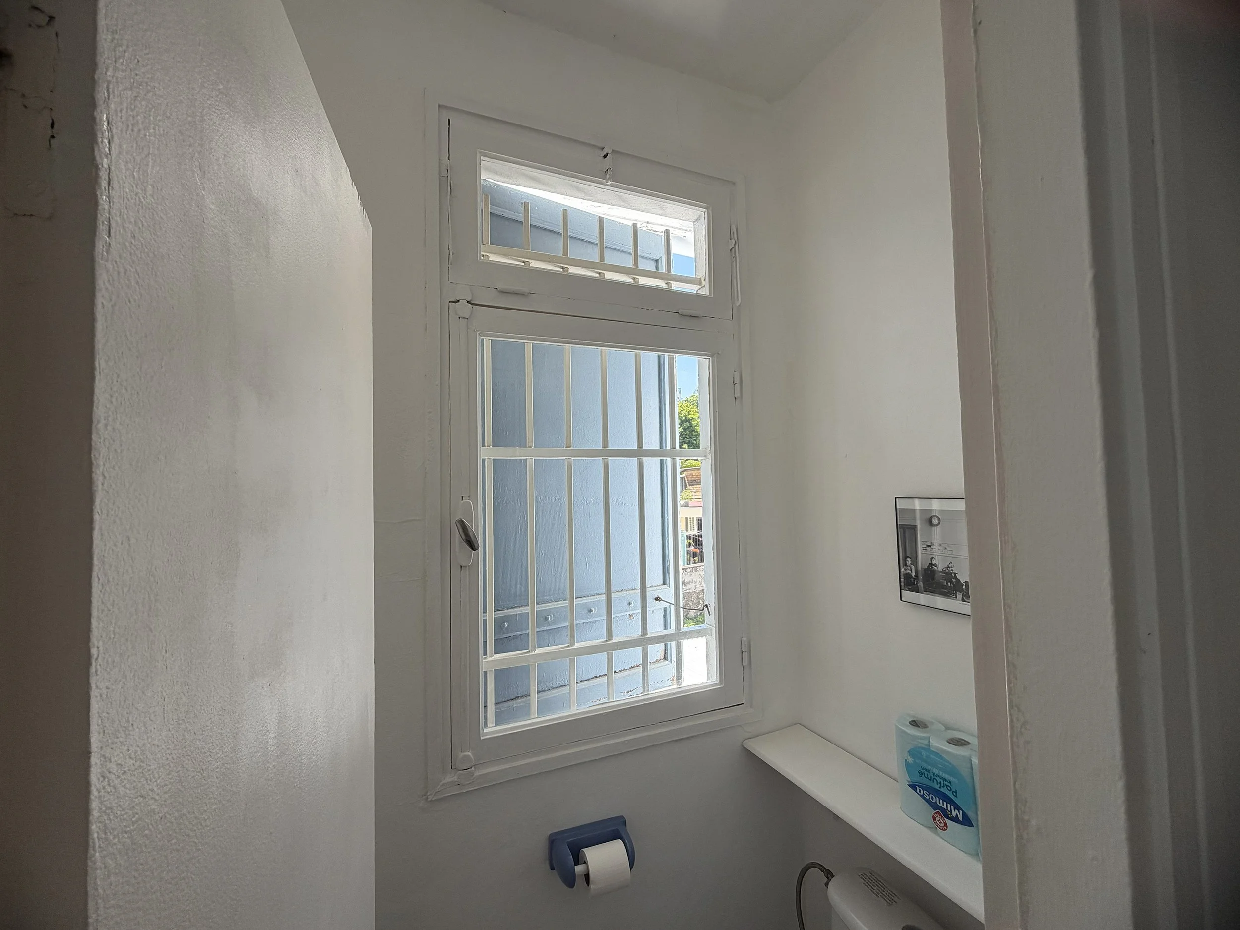 Small bathroom with a window, toilet paper roll, and soap holder with toilet, toiletries, and a picture on the wall.