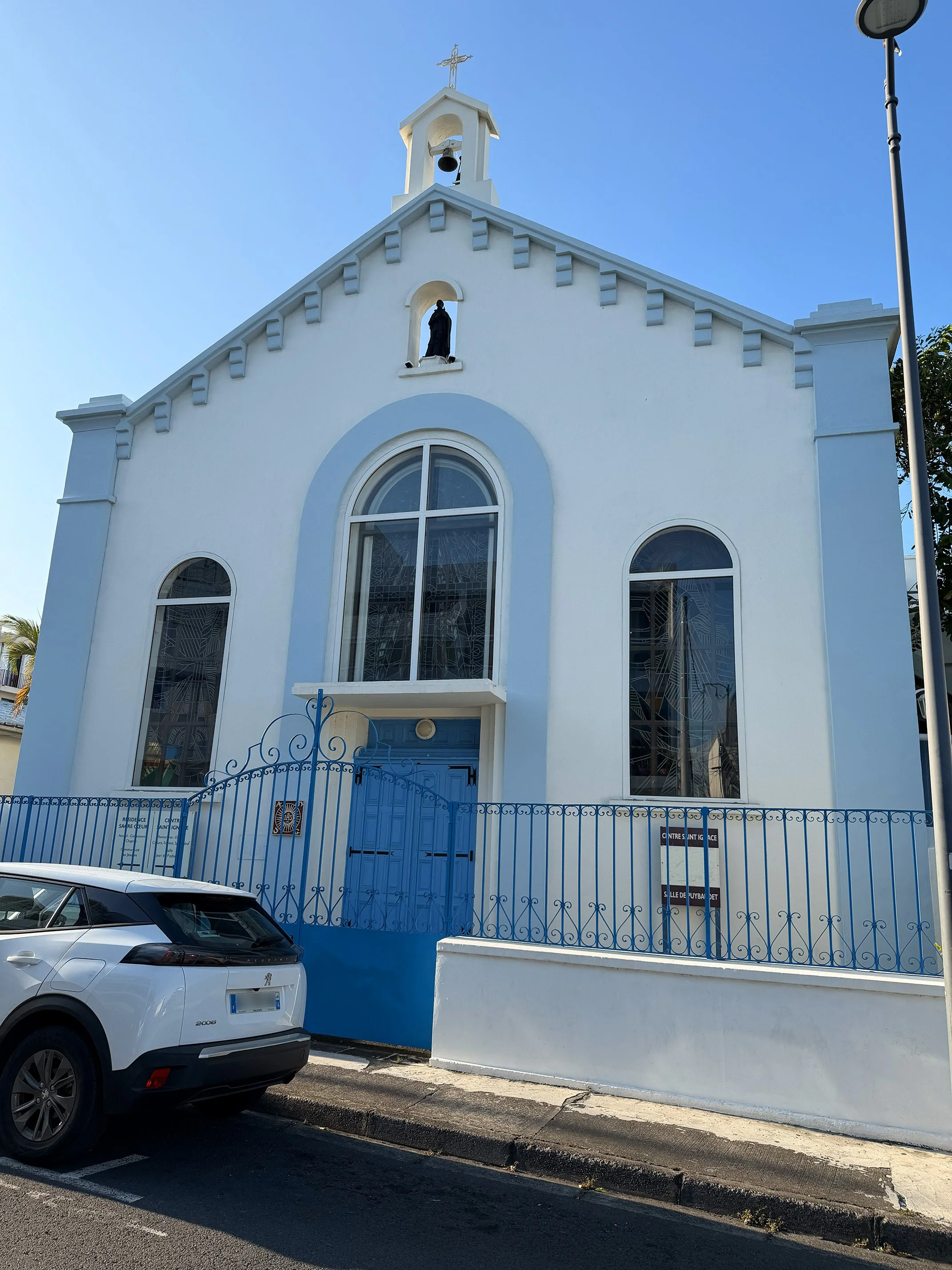 White church with blue accents, arched windows, a gated entrance, and a cross on top, parked along the street on a sunny day.