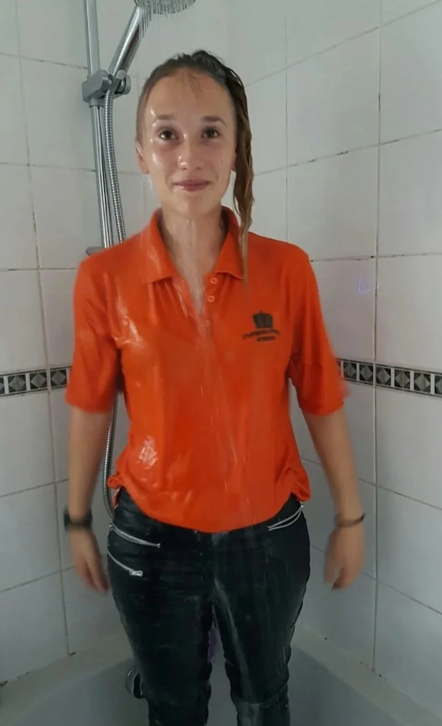 Young woman in orange shirt standing in a shower with water pouring on her.