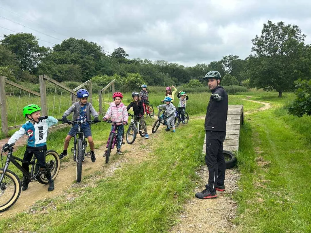 A group of children wearing helmets on bikes listening to an instructor on a grassy outdoor trail.