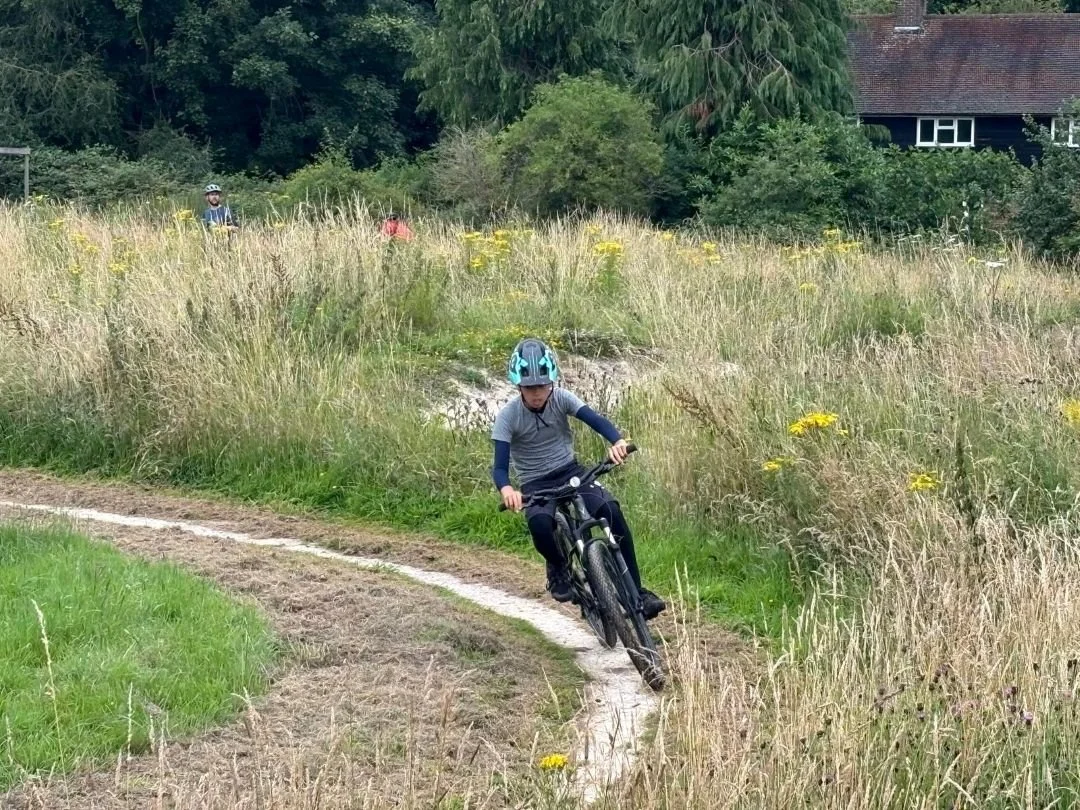 A child riding a mountain bike at a Muddy MTB Session on a narrow dirt trail through tall grass and wildflowers, with two other bike riders and a house with trees in the background.