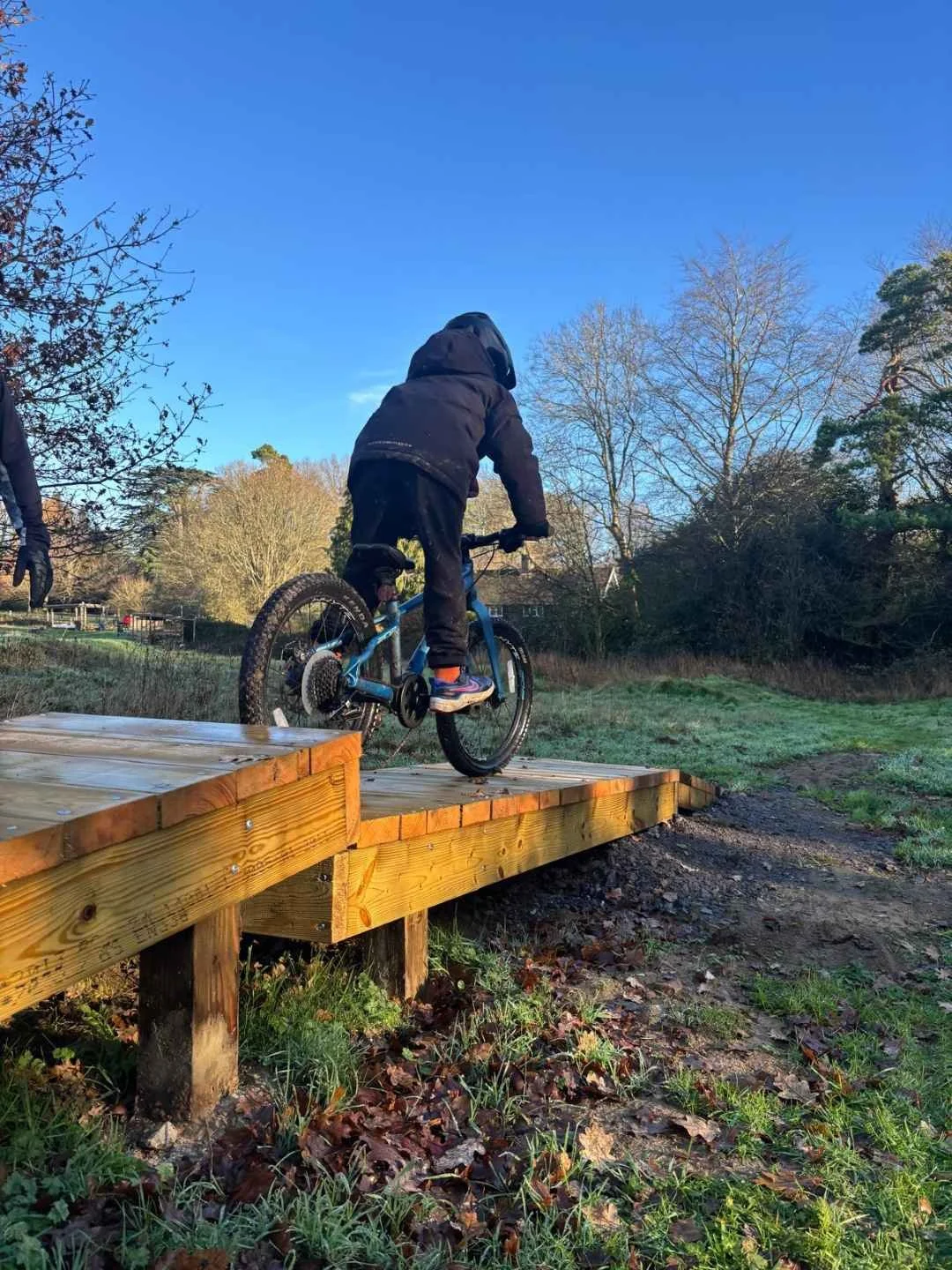 Child riding a mountain bike on a wooden ramp in an outdoor park during fall, with bare trees and clear blue sky in the background.