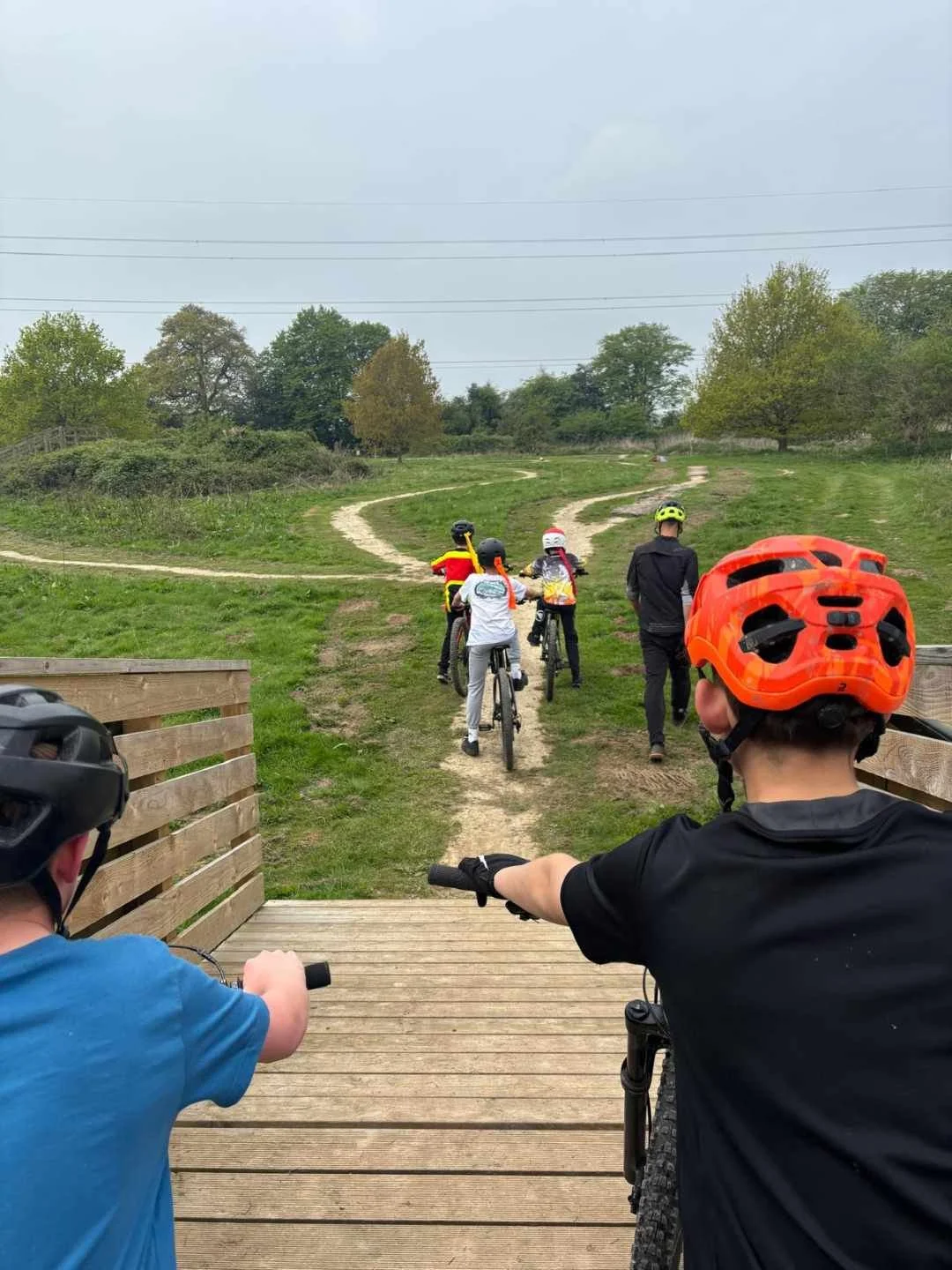 Group of children and adults riding bikes on a dirt trail through a grassy park, wearing helmets, with cloudy sky overhead.
