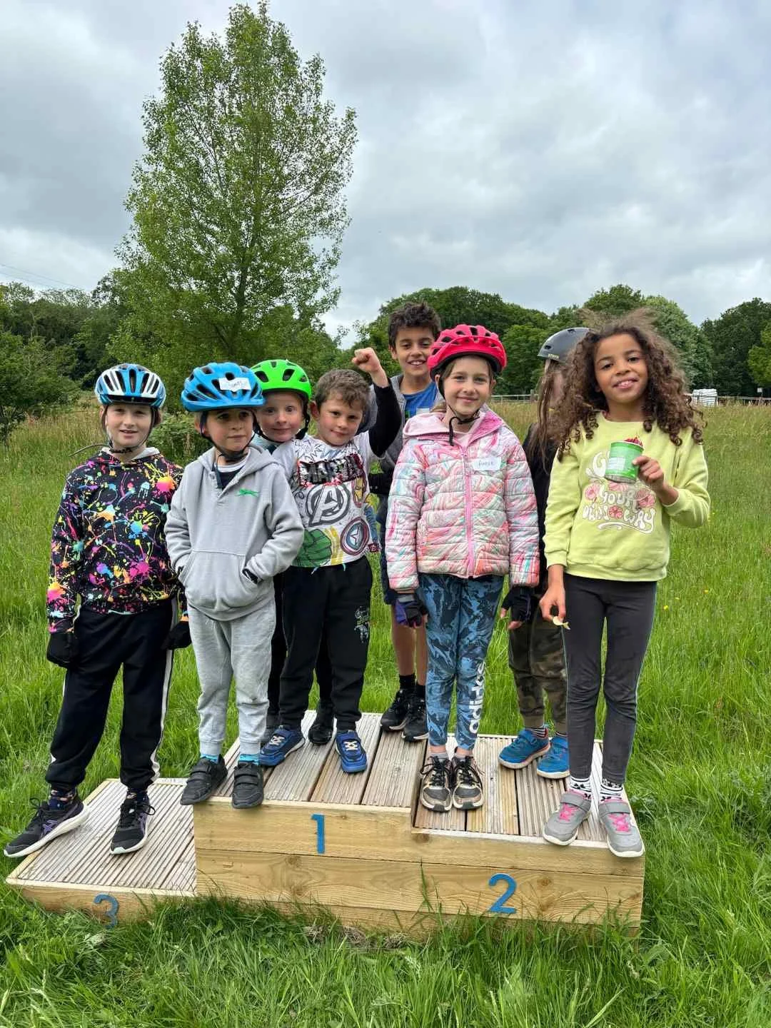 Group of children wearing helmets standing on a winners' podium outdoors on a grassy field with trees and overcast sky in the background.