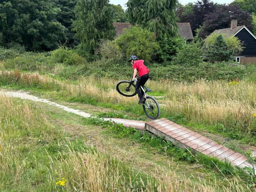 Person wearing a red jacket and a black helmet performing a jump on a bicycle on a small wooden ramp in a grassy outdoor area.