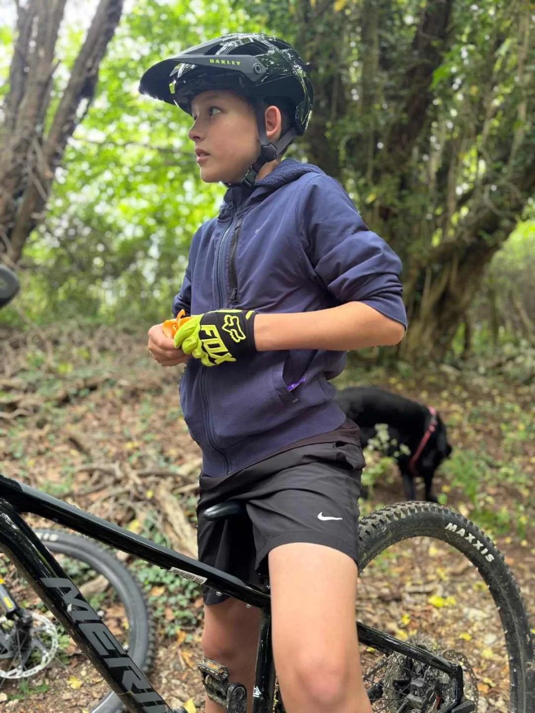 Young boy wearing a black helmet, blue jacket, and black shorts, standing with his mountain bike in a wooded outdoor setting. He has yellow and black gloves and is holding an orange object. A black dog is in the background near trees.