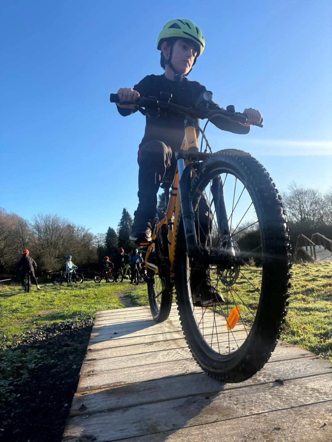 Child wearing a yellow helmet riding a mountain bike on a wooden track, with a group of children and adults on bikes in the background on a sunny day.
