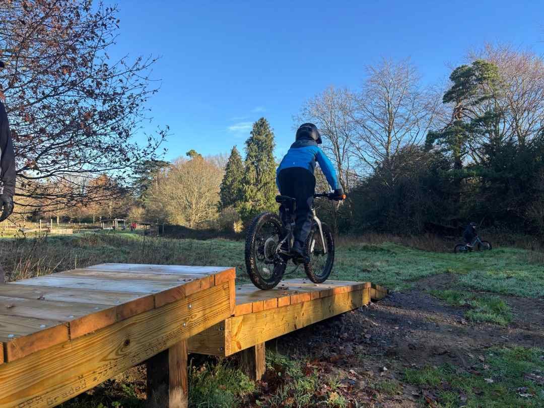 A person in a blue jacket and black helmet riding a mountain bike off a wooden ramp outdoors on a clear day with trees in the background.