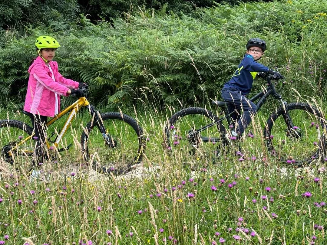 Two children wearing helmets riding mountain bikes on a trail through tall grass and wildflowers, with lush green foliage in the background.
