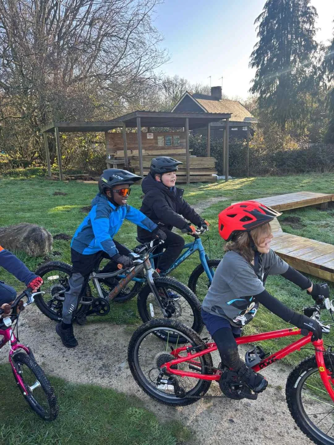 Three children riding bicycles outdoors on a sunny day, wearing helmets and appropriate gear, on a grass and dirt path surrounded by trees and a house in the background.