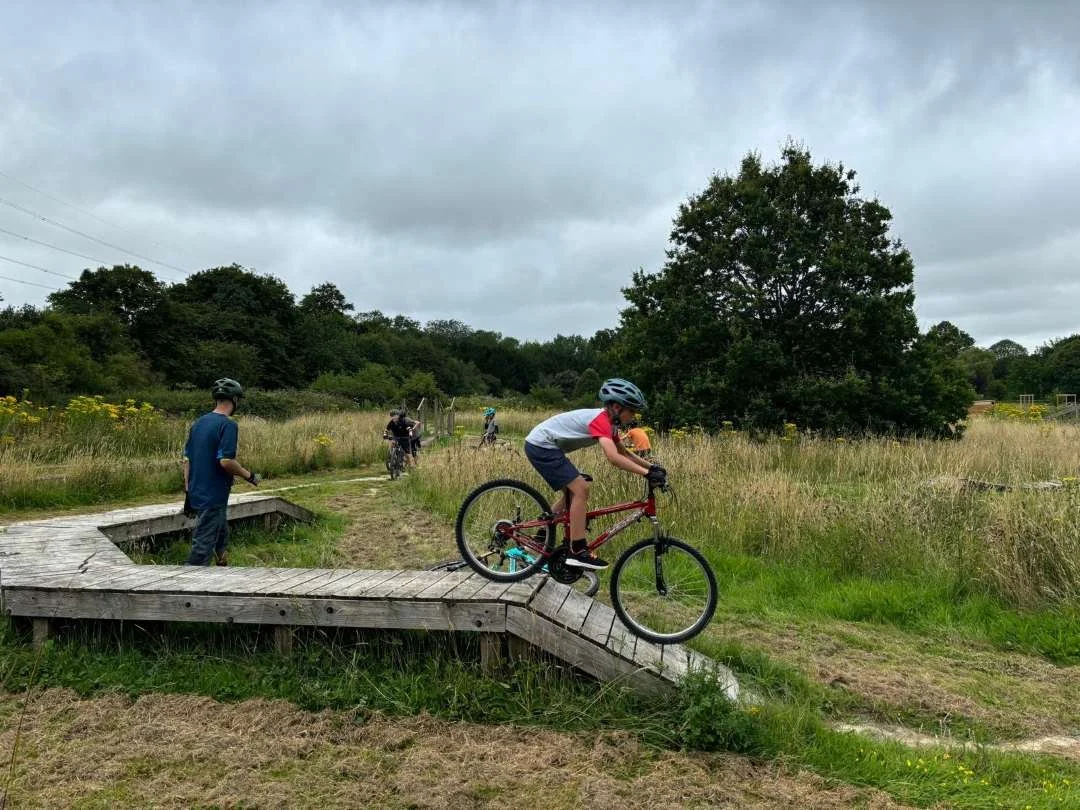 A boy in a helmet riding a mountain bike down a wooden ramp in a grassy outdoor area, with other children and adults in the background under cloudy skies.