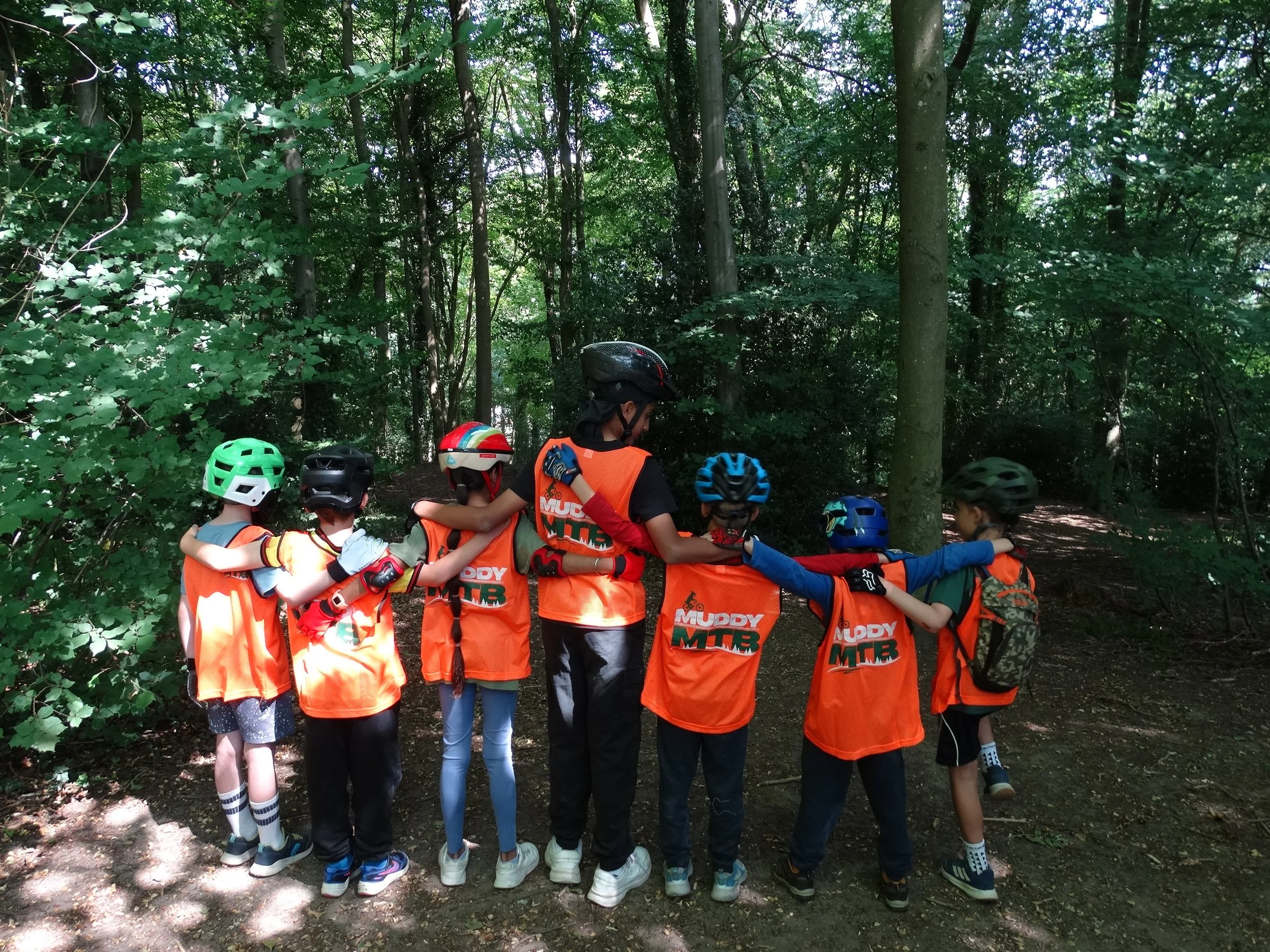 Group of children wearing helmets standing on a winners' podium outdoors on a grassy field with trees and overcast sky in the background.