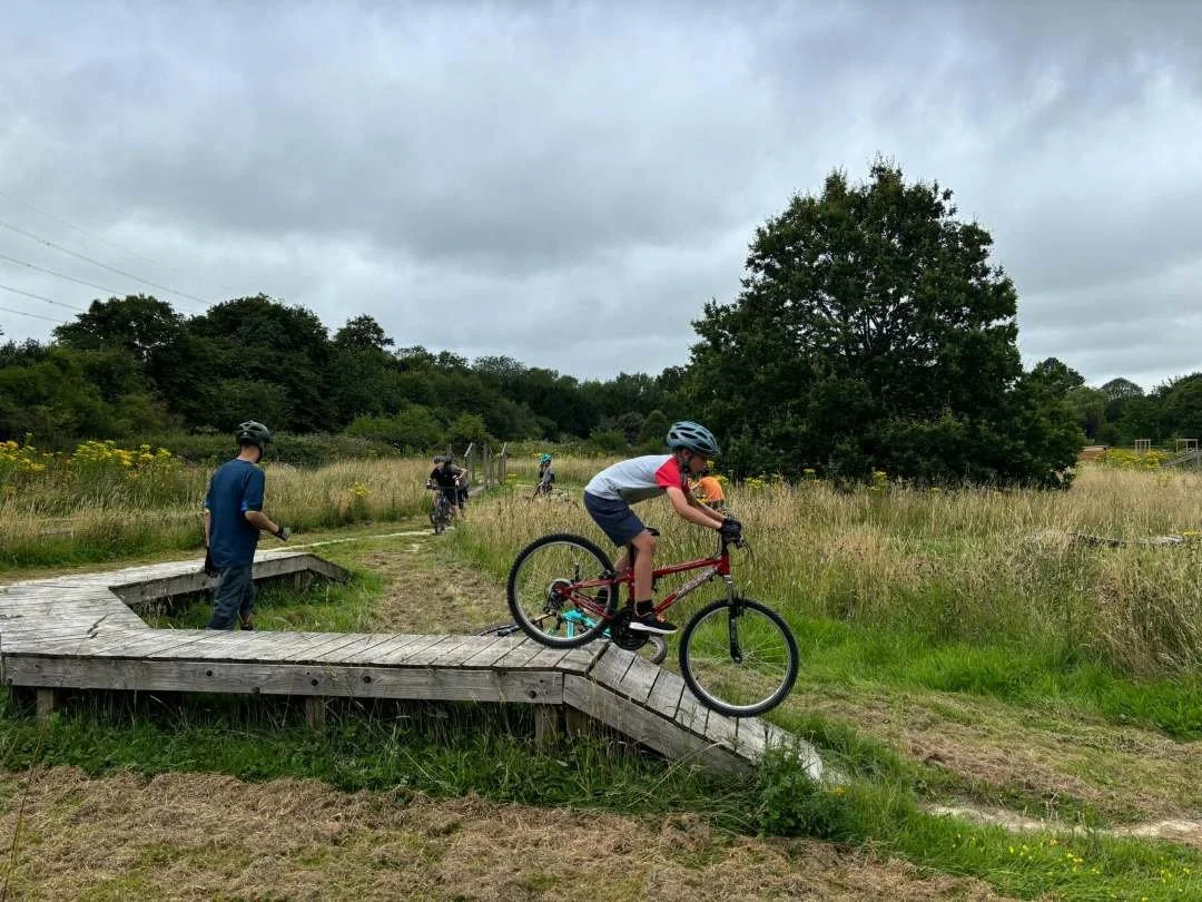 A child in a helmet riding a mountain bike down a wooden ramp on a dirt trail in a grassy park, with other kids and adults in the background under a cloudy sky.