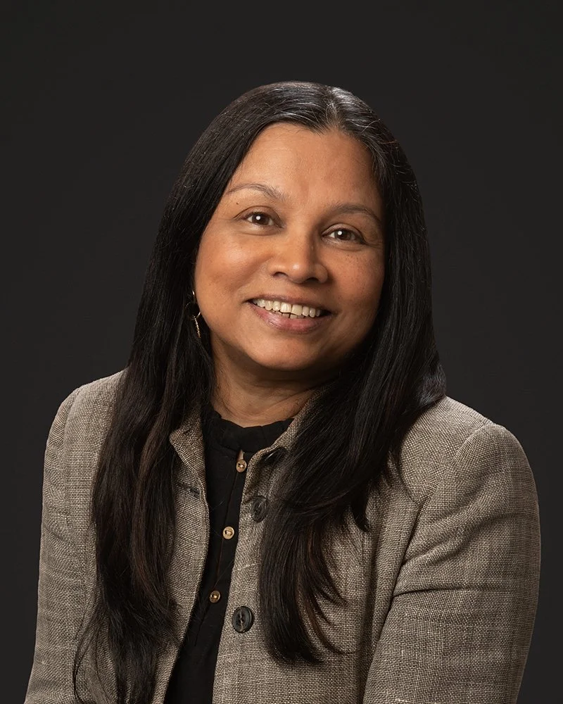 Portrait of a woman with long black hair, wearing a gray blazer and black top, smiling against a dark background.