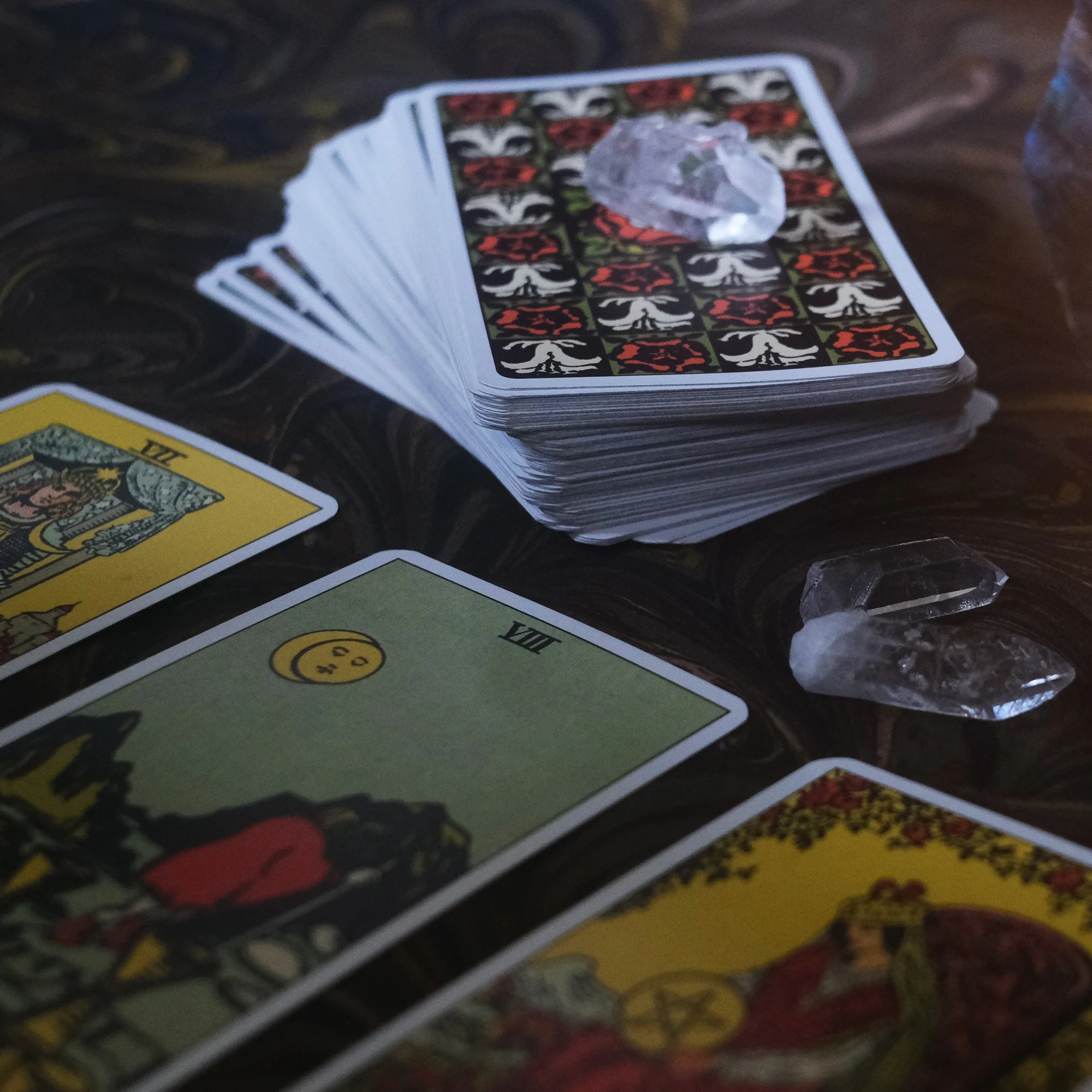 A spread of tarot cards on a dark patterned surface with two crystals nearby, and a deck of cards with a crystal placed on top.