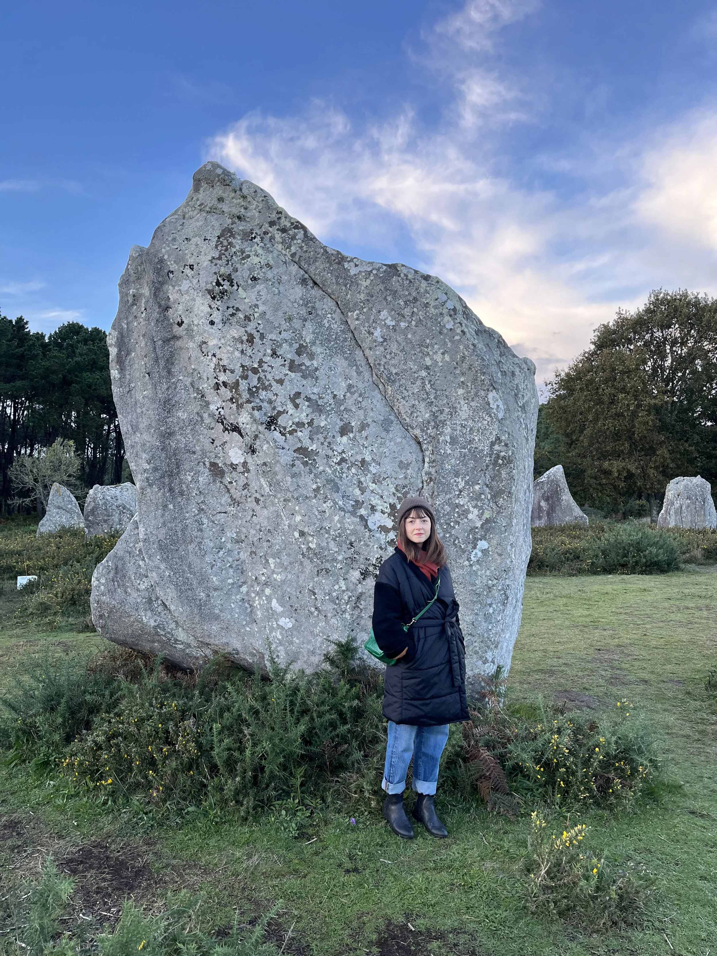A woman in a black coat, jeans, and boots standing next to a large boulder in an outdoor park or field with trees and a partly cloudy sky in the background.