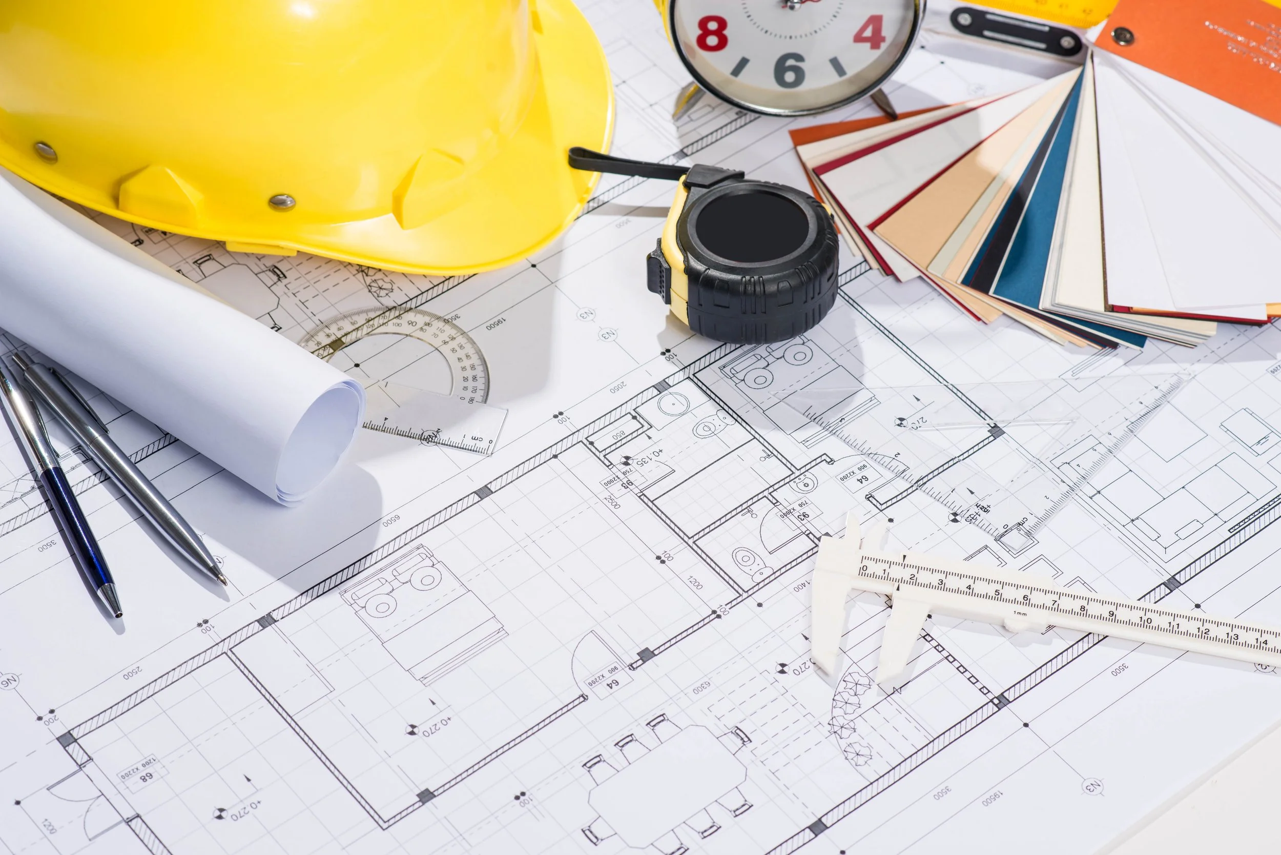 Architectural blueprints and tools, including a yellow hard hat, a measuring tape, a protractor, and a fan deck of paint colors, arranged on a table.