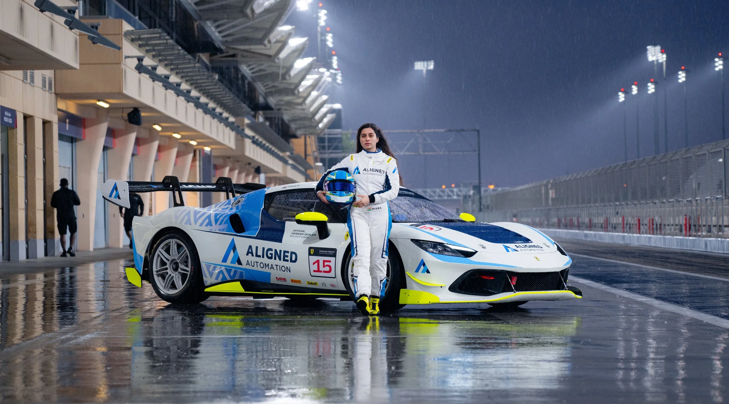 Diana Pundole standing next to a race car on a wet race track at night, holding a helmet. The race car is white with blue and yellow accents and has the number 15. The driver is wearing a white racing suit with sponsor logos.