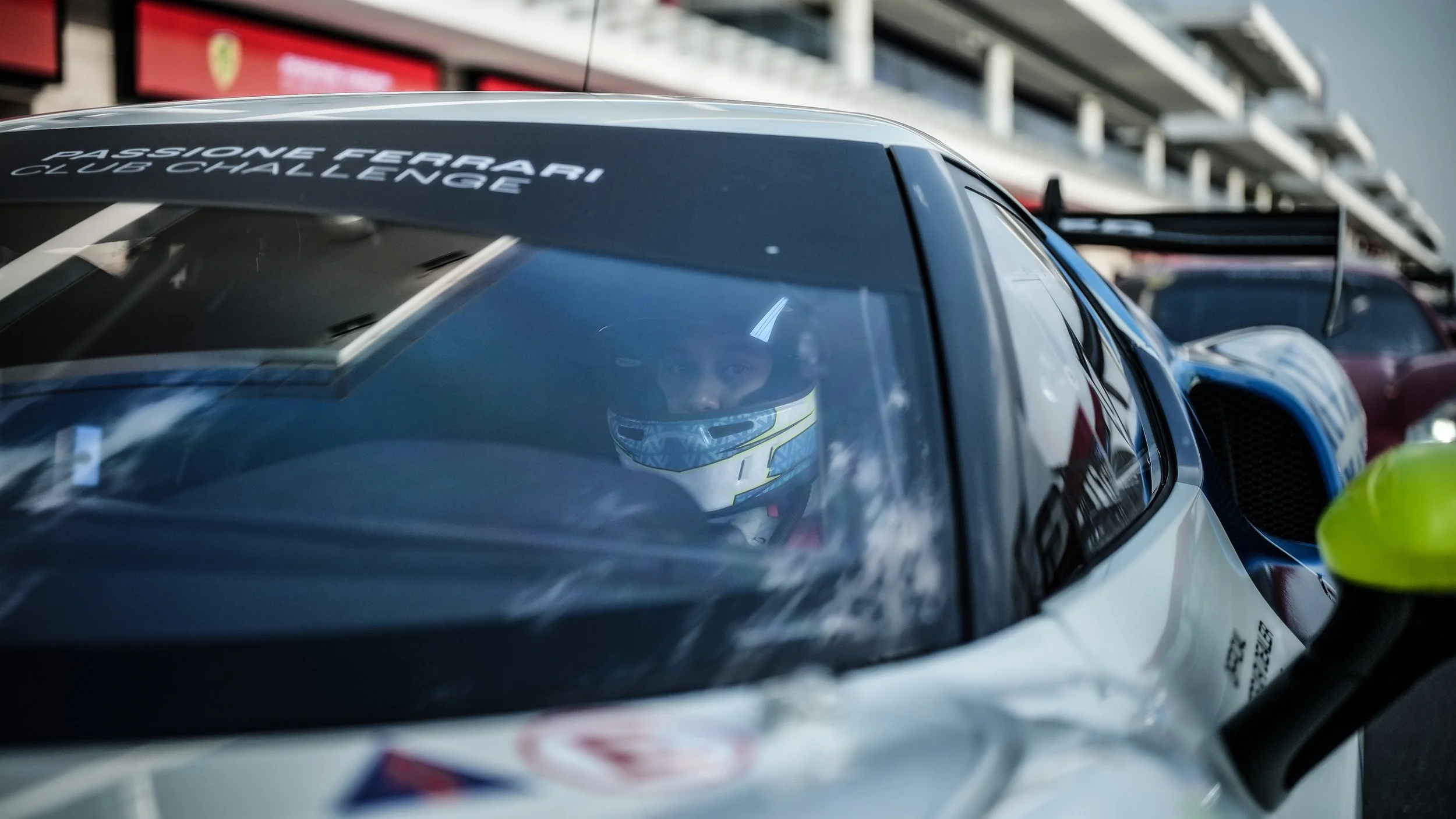 Close-up of a racing car with Diana Pundole wearing a helmet inside, parked on the track with a blurred background of grandstands and other cars.