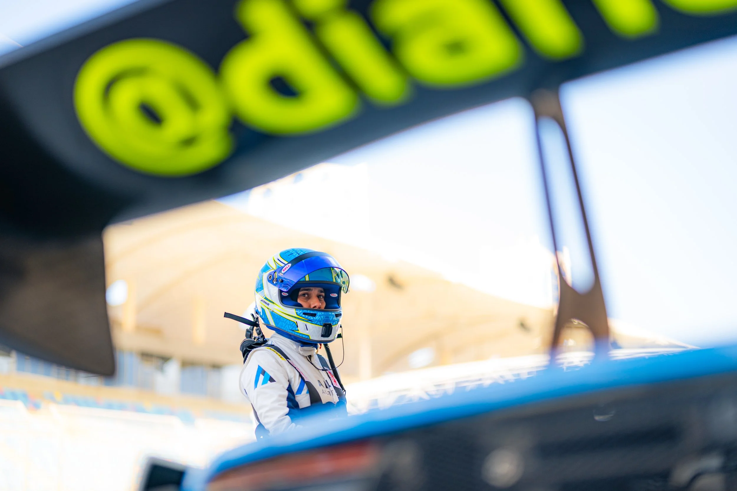 Diana Pundole wearing a blue and yellow helmet, standing beside a race car, with the photo viewed through parts of the car's spoiler or body in the foreground.