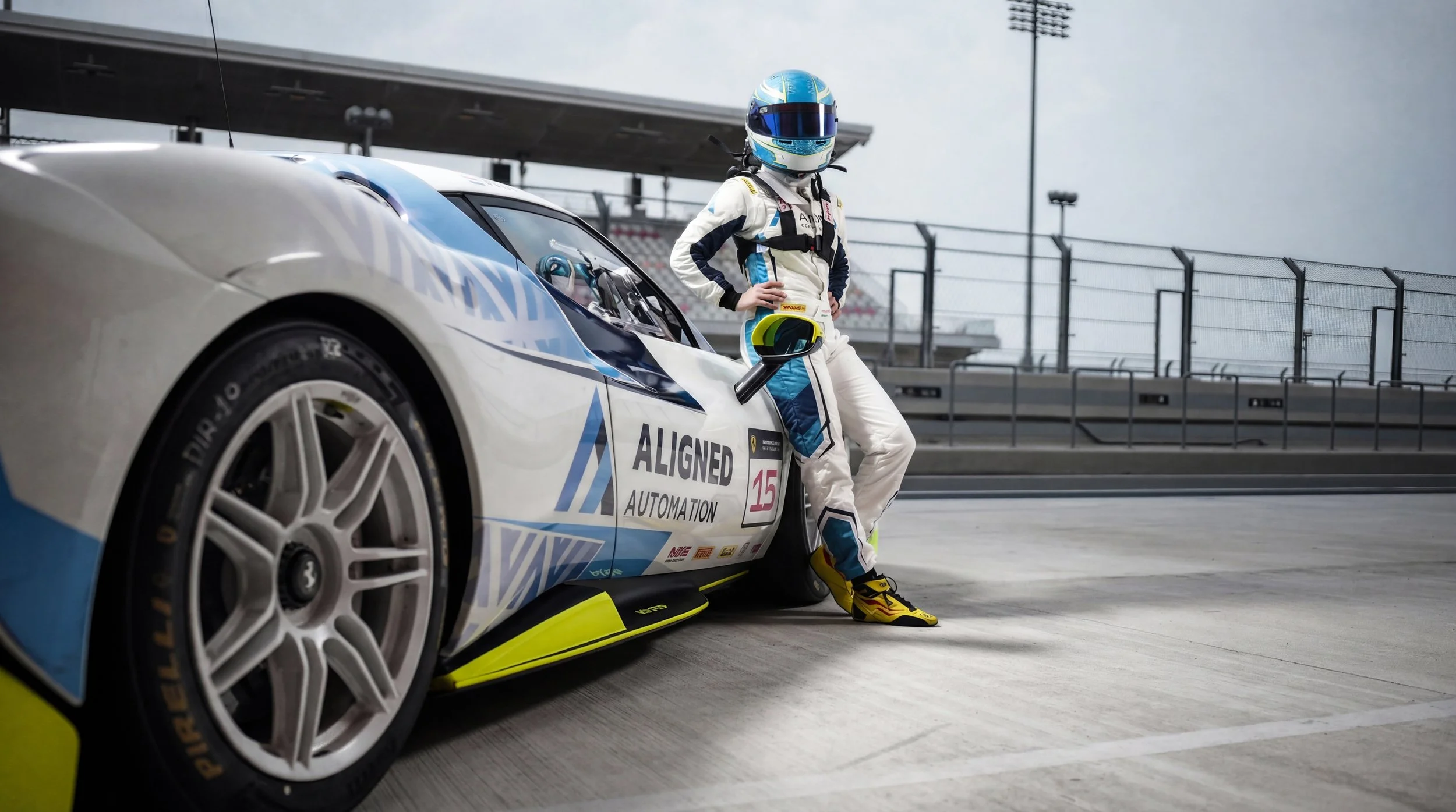 Diana Pundole in a white and blue racing suit with a blue helmet, standing next to a white race car with blue accents on a racetrack.