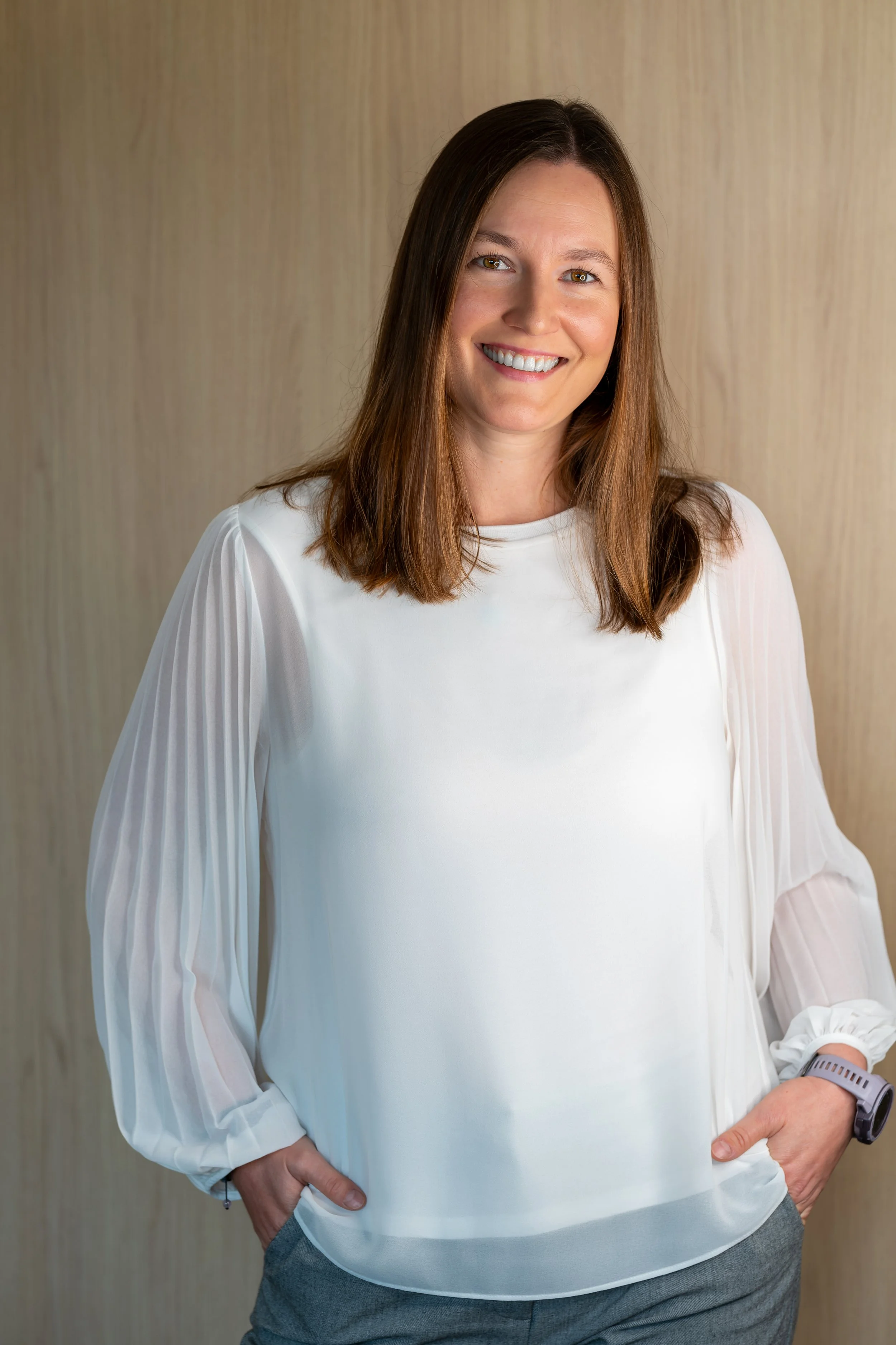A woman with shoulder-length brown hair smiling, wearing a white blouse with pleated sleeves and gray pants, standing against a wooden background.