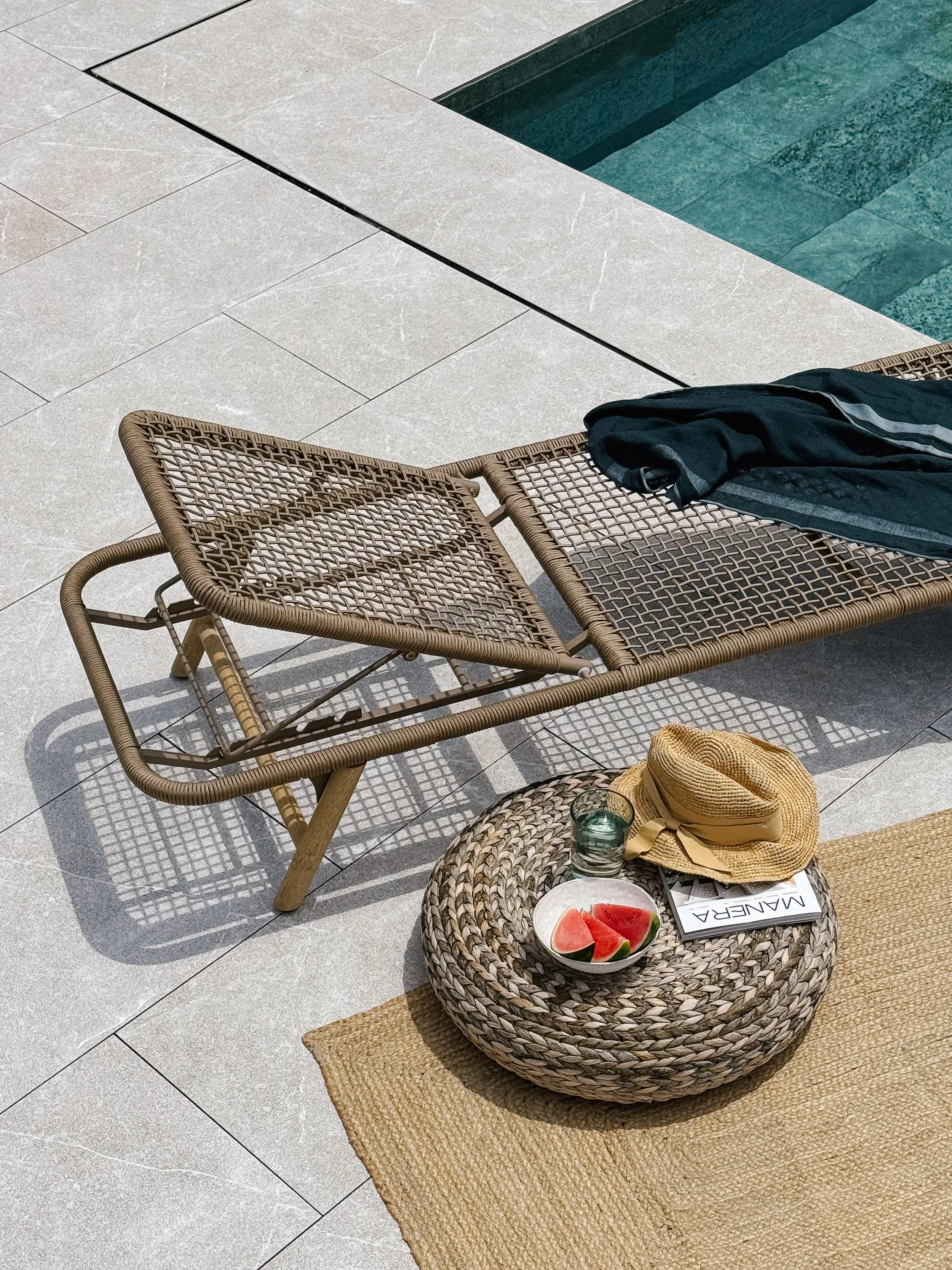 A poolside scene with a woven lounge chair, a round woven ottoman with watermelon slices, a hat, a glass of water, a magazine, and a towel, with a swimming pool in the background.