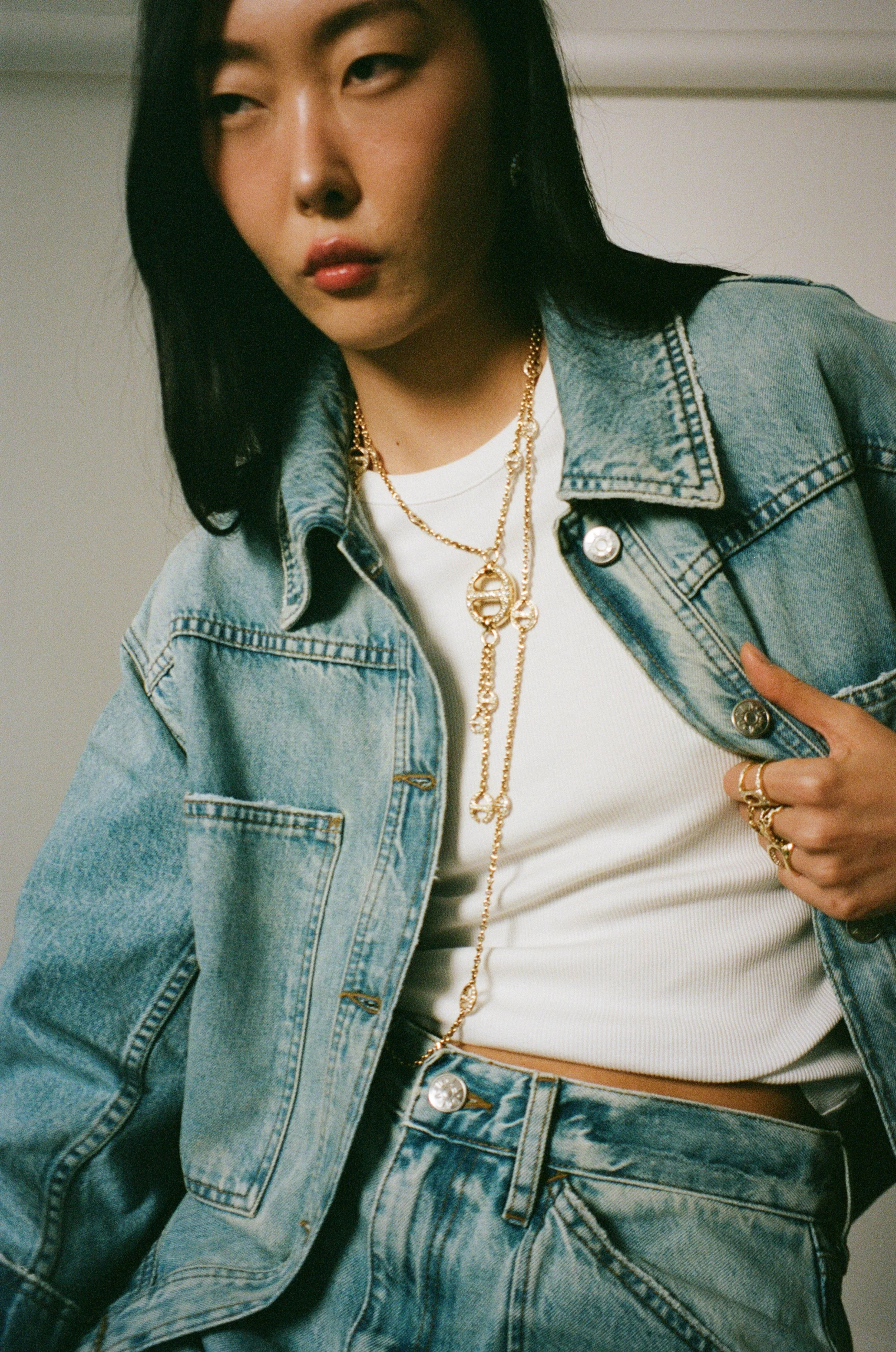 A woman wearing a denim jacket, white crop top, multiple gold necklaces, and rings, with black hair partially covering her face.