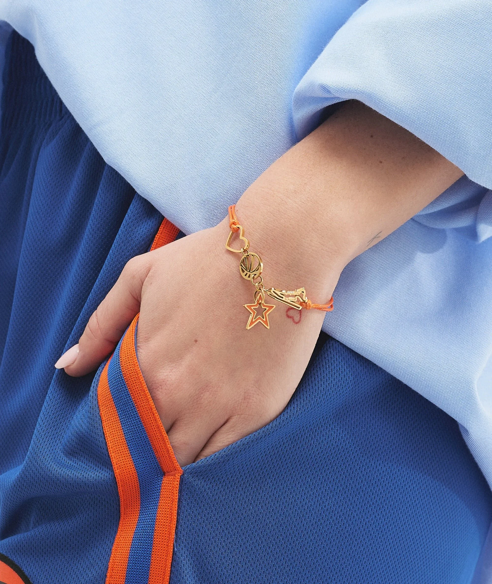Close-up of a person's hand and wrist, wearing a blue sports jacket with orange and blue striped trim, and a gold charm bracelet with various charms including a star and heart.