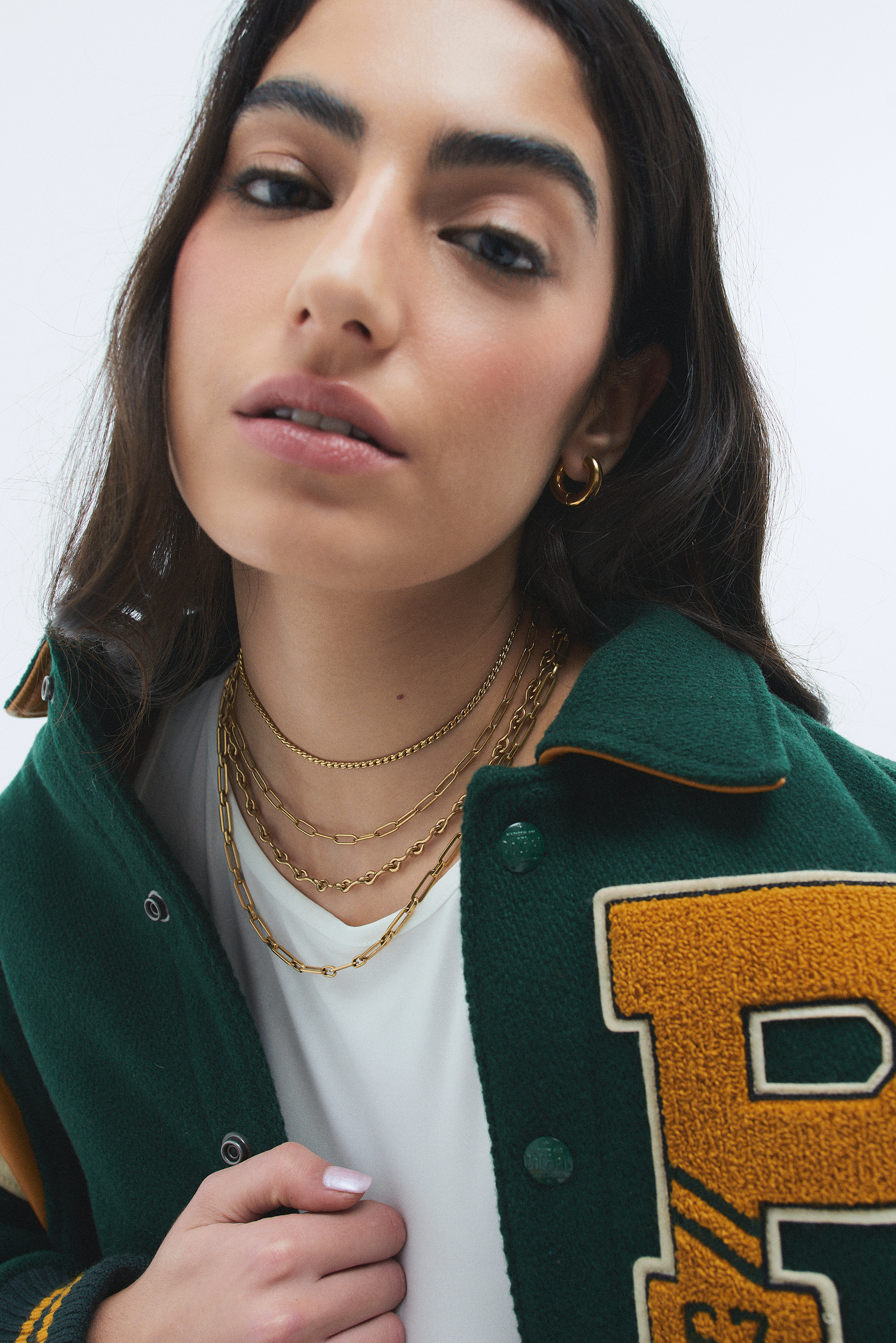 Close-up of a young woman with brown hair, wearing gold jewelry, a white shirt, and a green varsity jacket with a yellow letter patch.