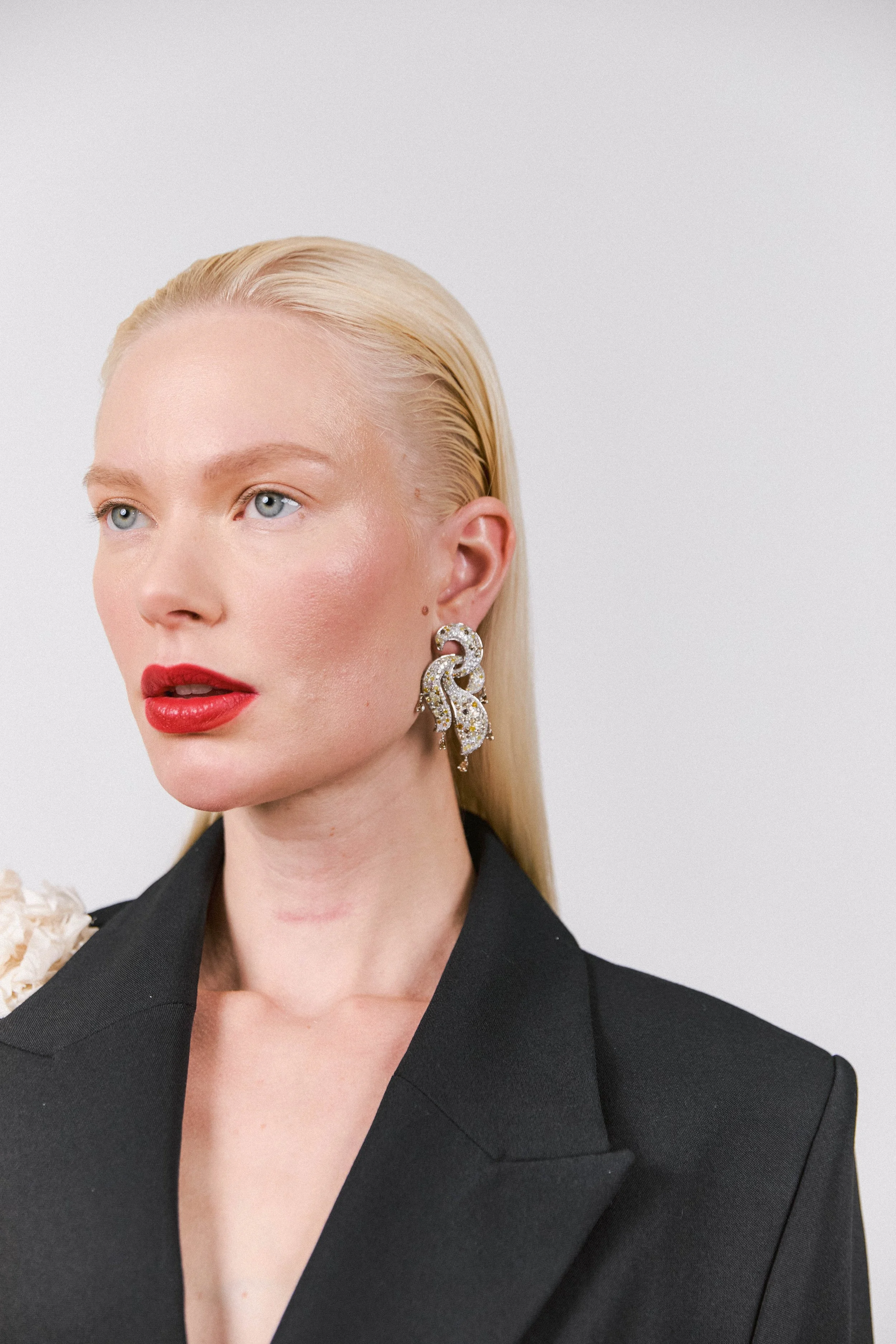 A woman with platinum blonde hair, red lipstick, wearing large ornate earrings, and a black blazer against a plain white background.