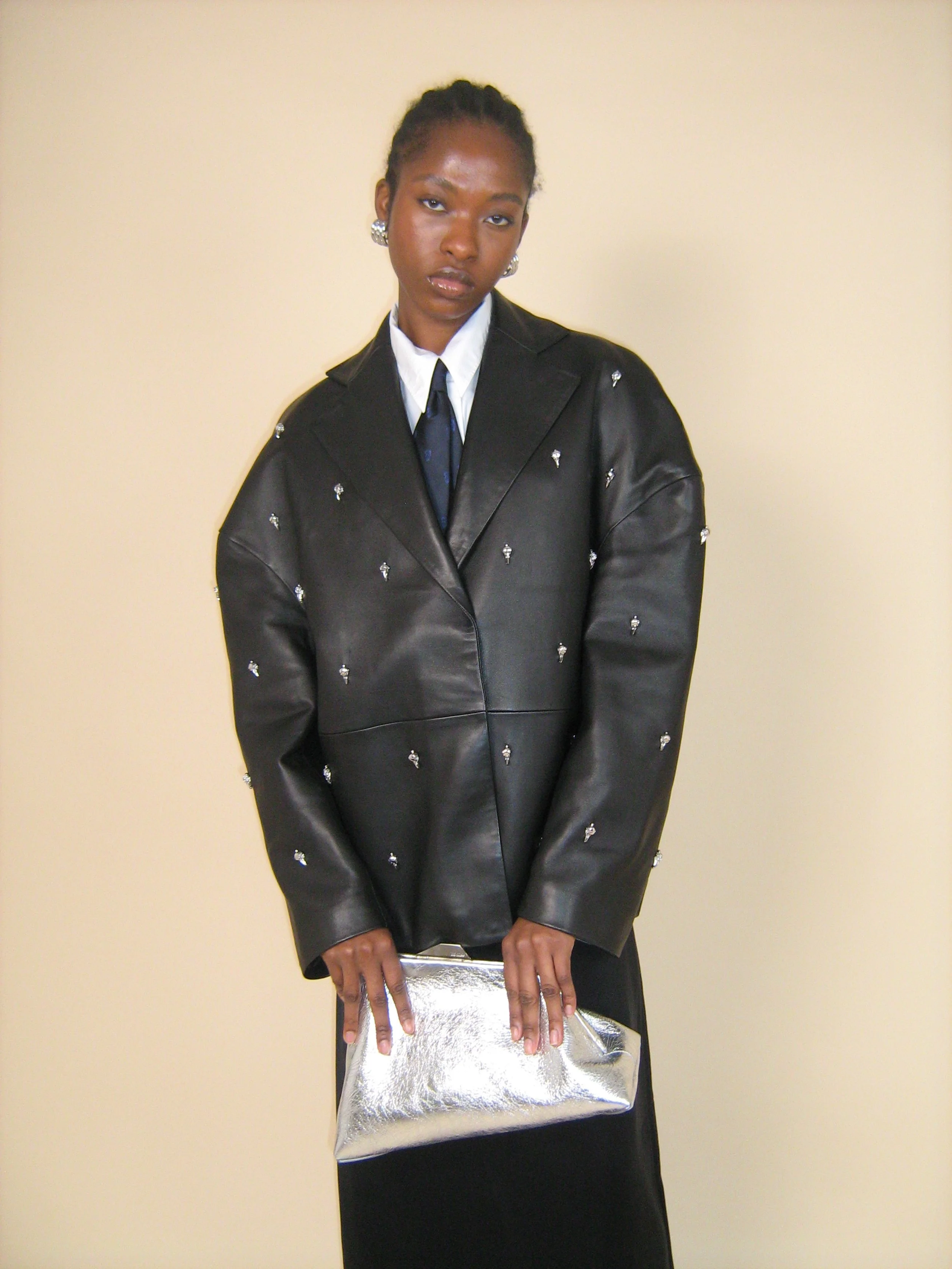 Young woman in black leather jacket with small embellishments, white shirt, black tie, black skirt, holding a metallic silver clutch, standing against a plain beige wall.