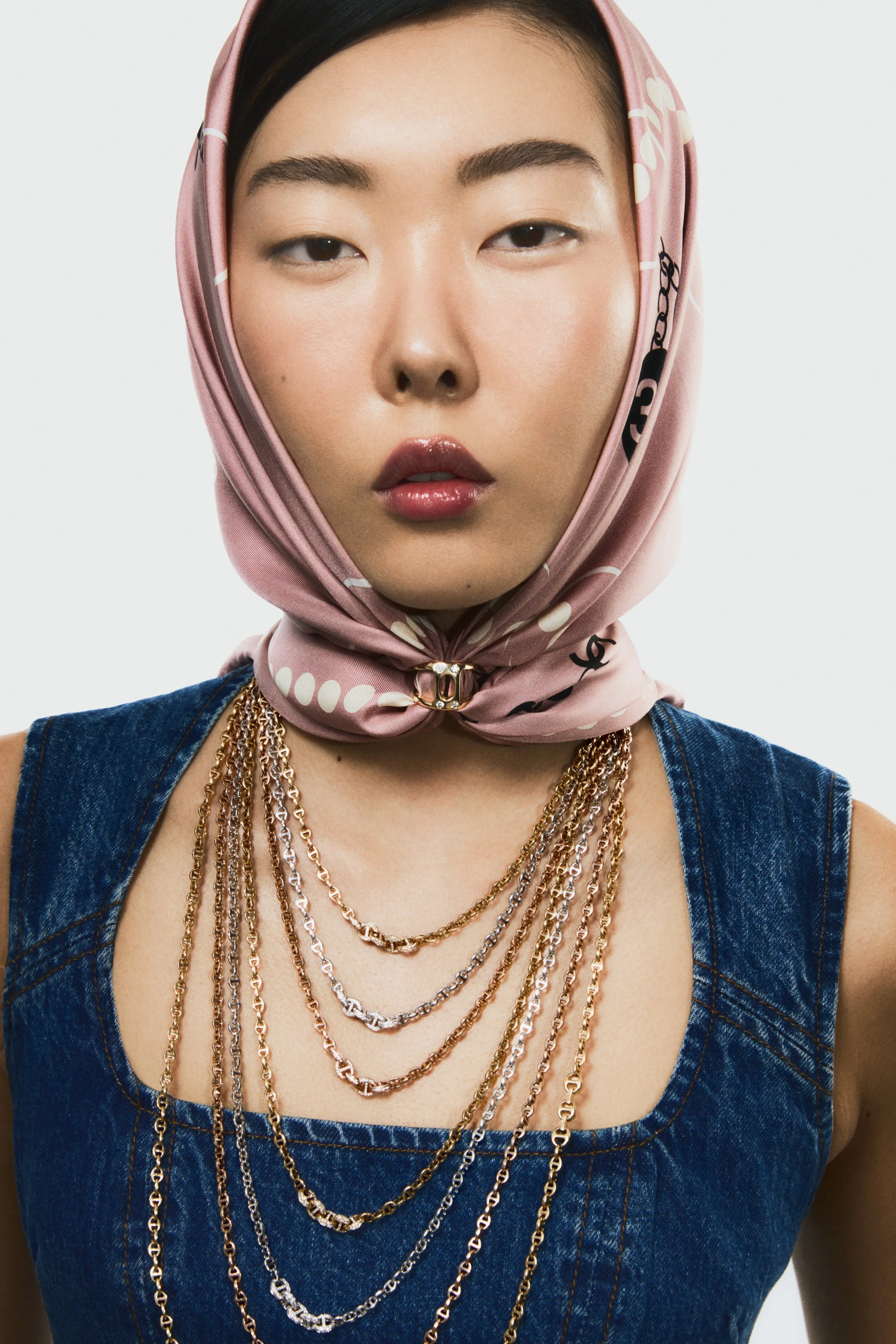 A woman wearing a pink scarf with chain detail and a sleeveless denim top, accessorized with multiple layered gold and silver chains.