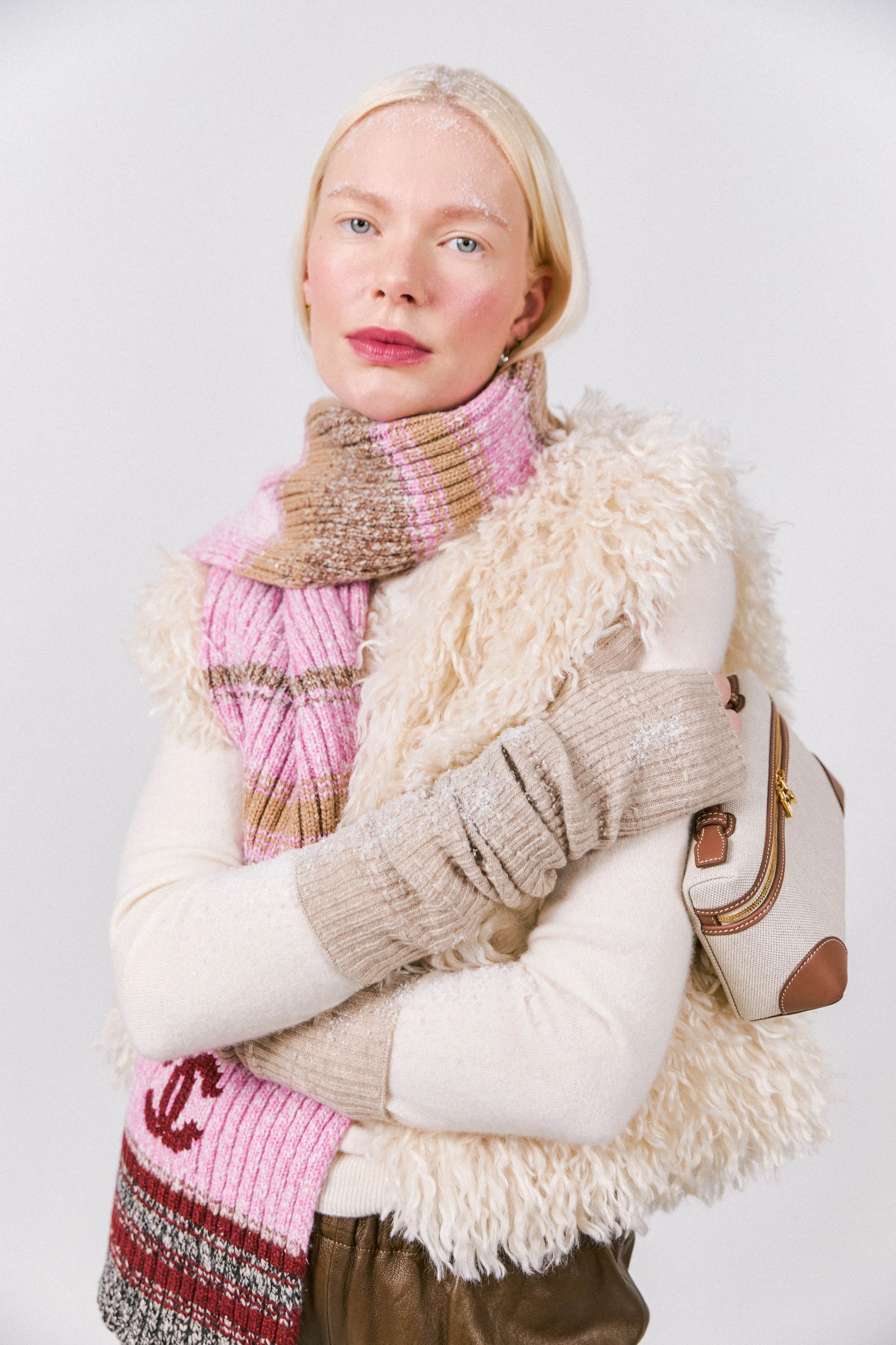 A woman with blonde hair and blue eyes wearing a beige sweater, a pink and brown striped scarf, a cream-colored fuzzy vest, and tan gloves, posing against a plain white background.