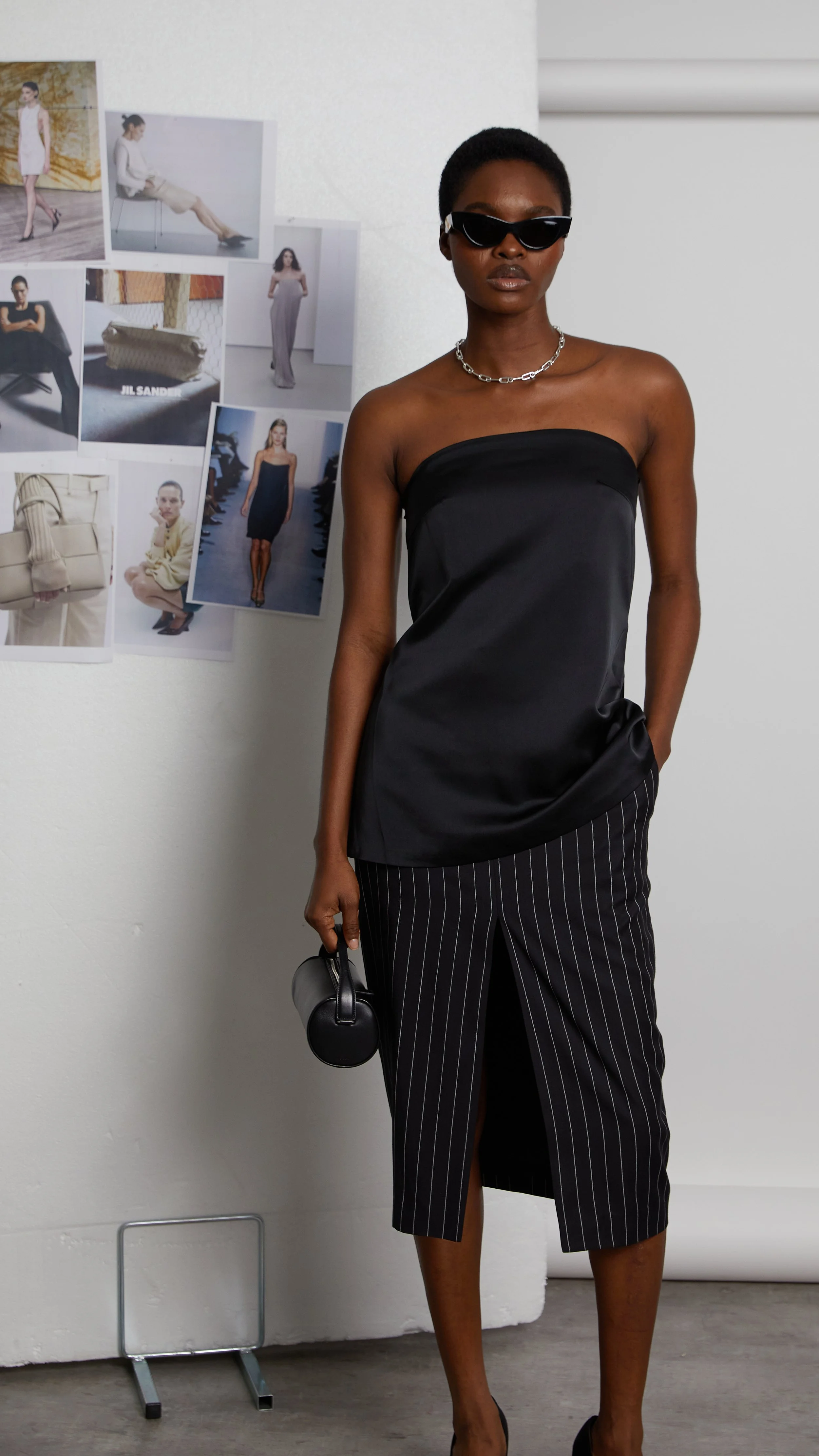 A stylish woman wearing black sunglasses, a strapless black top, pinstripe skirt with a front slit, holding a small black handbag, standing in front of a wall with fashion photos.