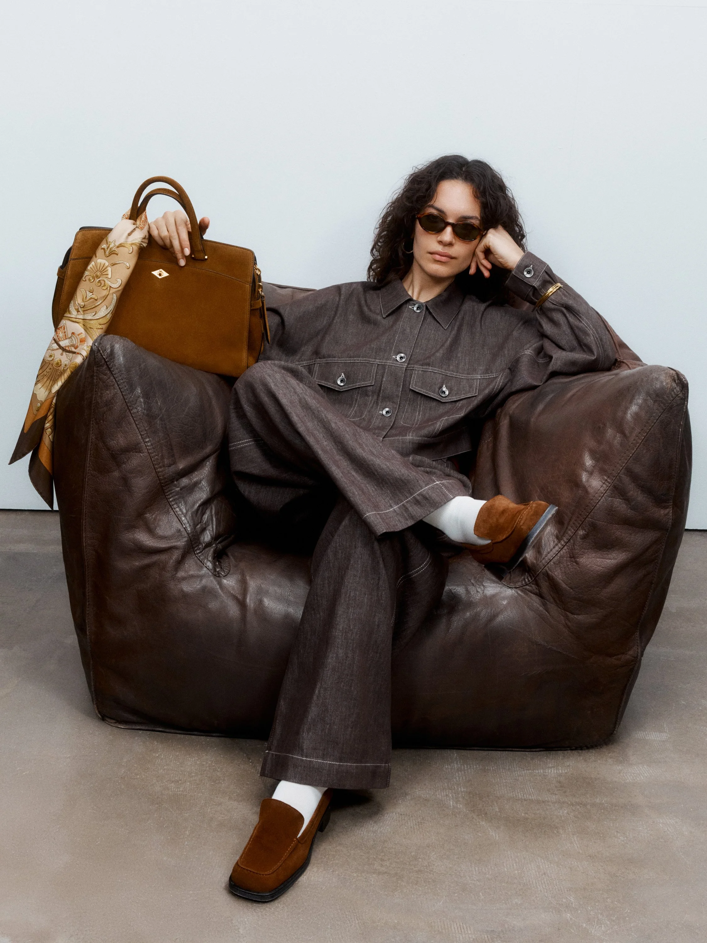 Woman sitting on a leather chair, wearing sunglasses, a brown jacket, brown pants, white socks, and brown shoes, with a brown handbag and a yellow scarf draped over the handbag.